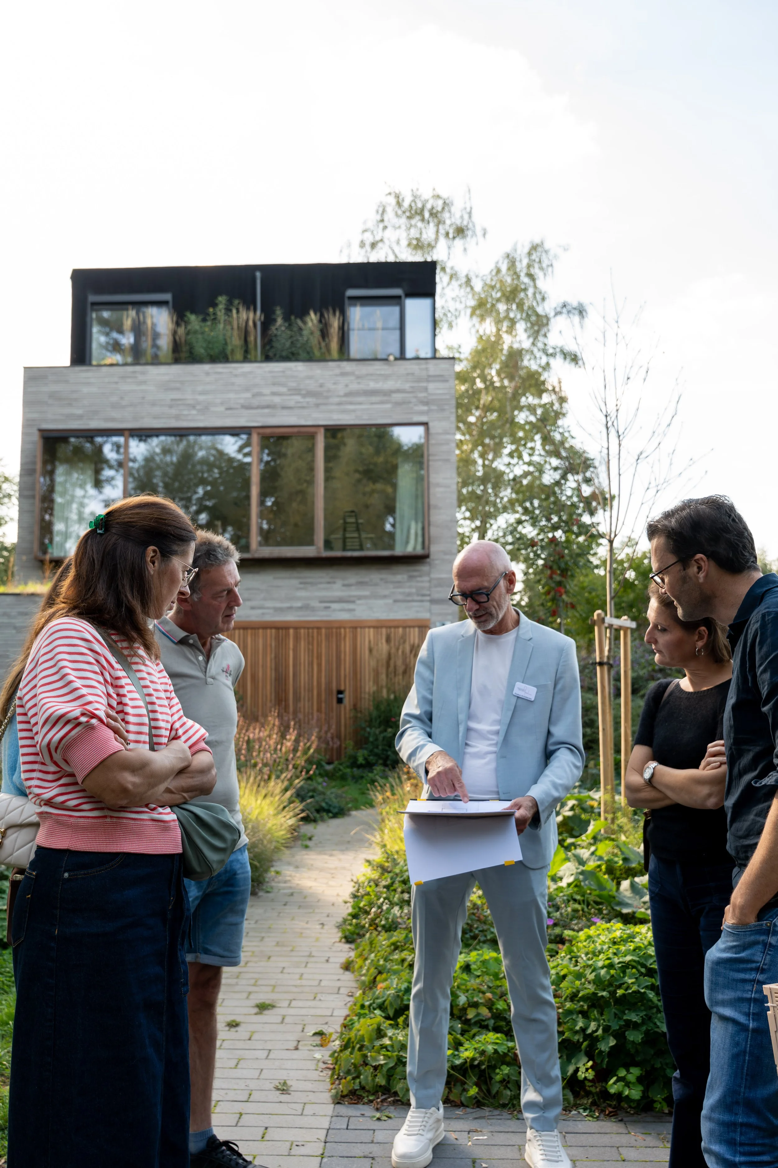 A group of five people gathered outdoors, listening to a man in a light blue suit who appears to be explaining something and holding a folded paper. They are in a garden or backyard area with a paved walkway, greenery, and a modern multi-story house in the background.
