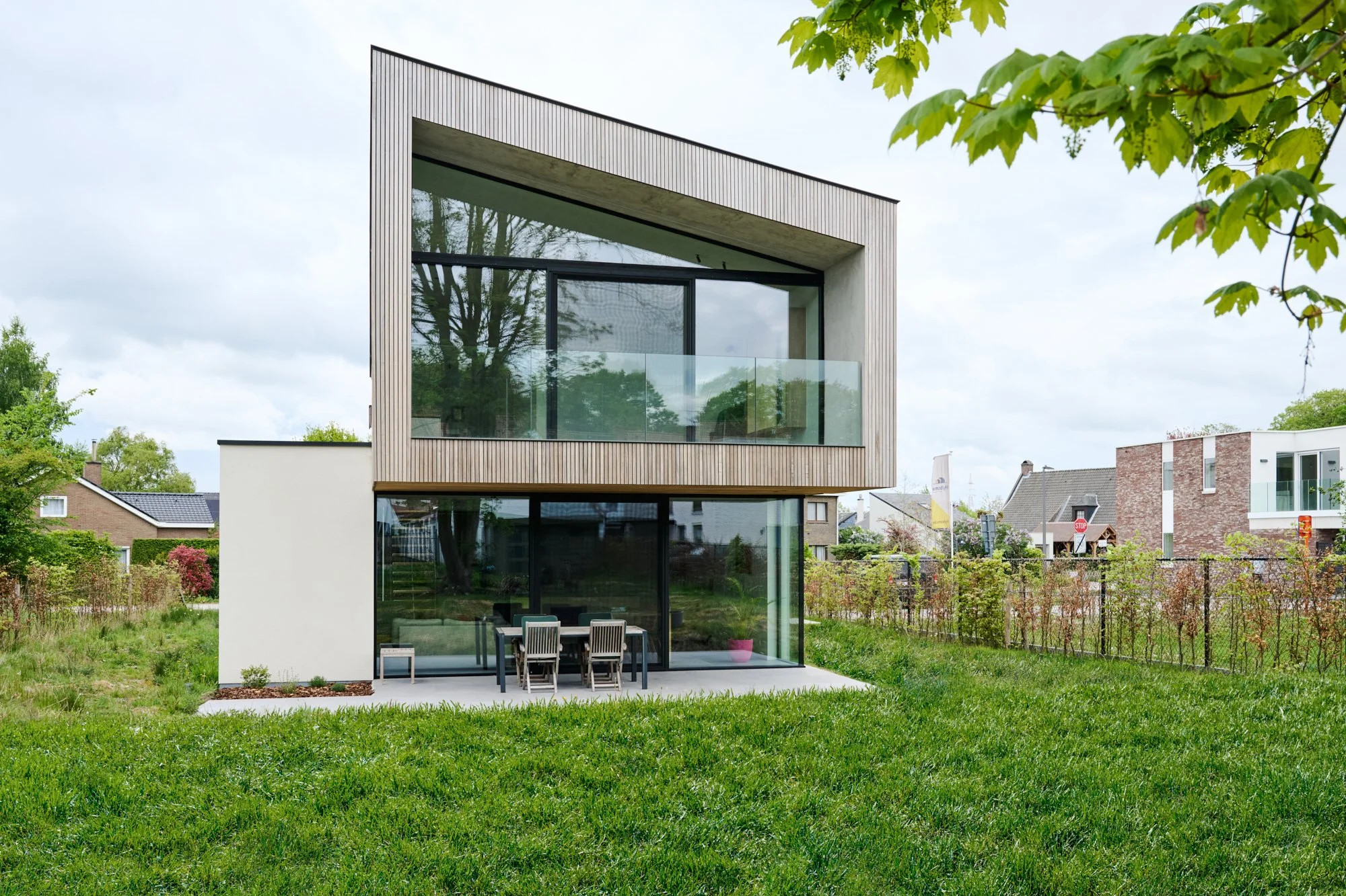 Modern two-story house with large glass windows and wooden exterior paneling, surrounded by green lawn and trees, in a suburban neighborhood.