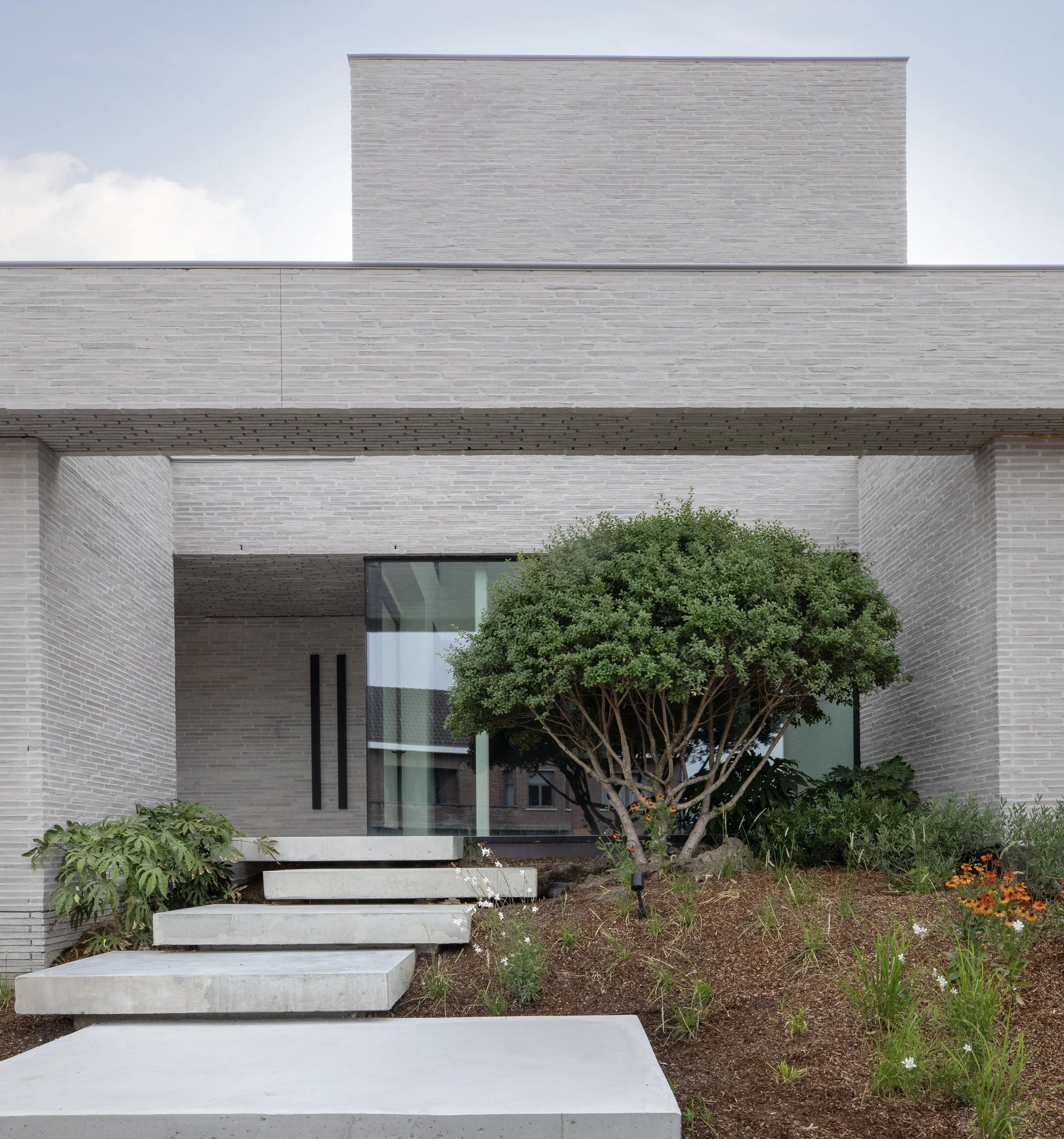 Modern house with gray brick exterior, glass windows, concrete steps, and landscaped garden with green shrub and flowers.