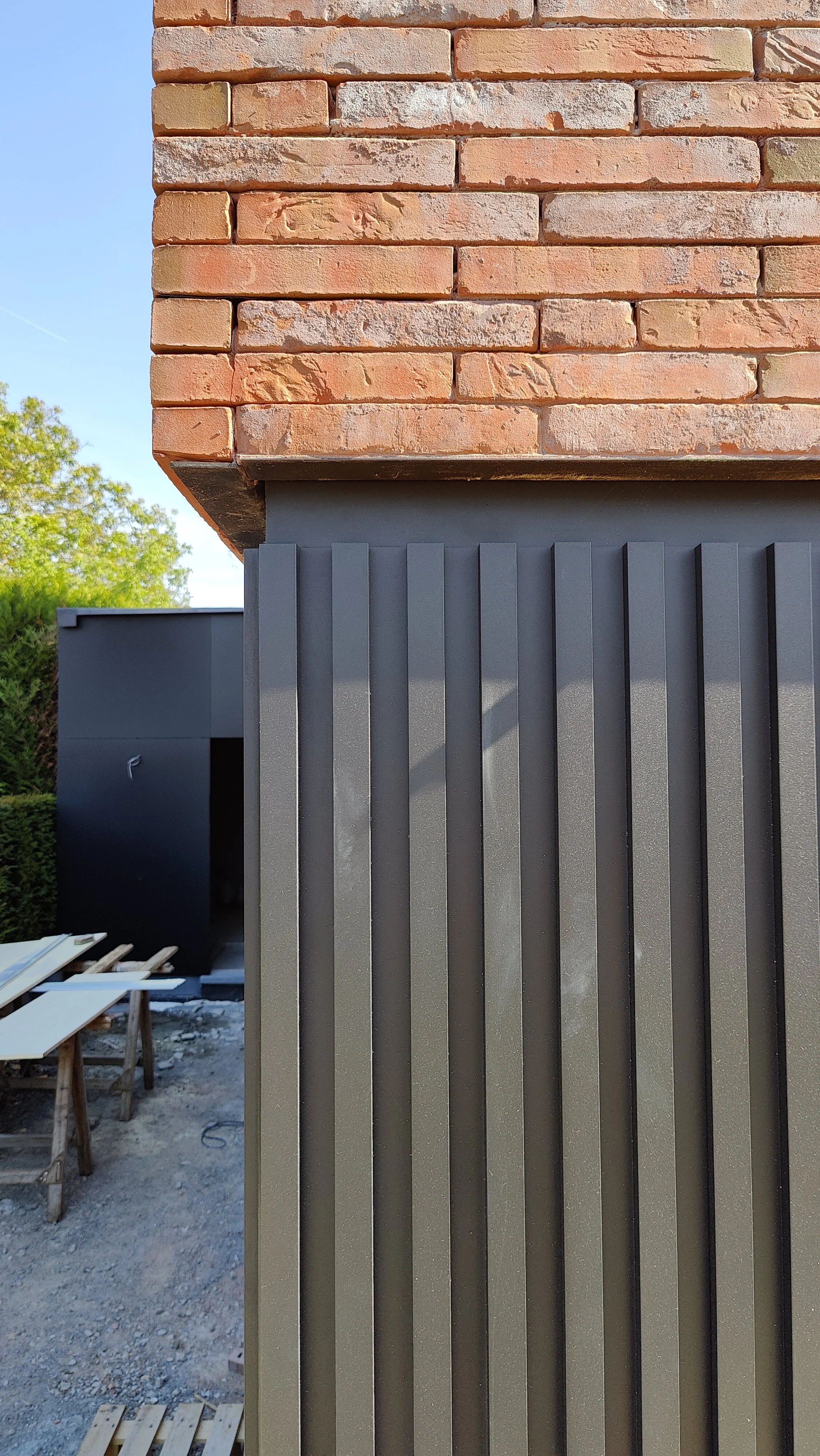 Close-up of a building corner showing red brick wall above dark gray vertically ridged metal panel siding, with a construction site and trees in the background.