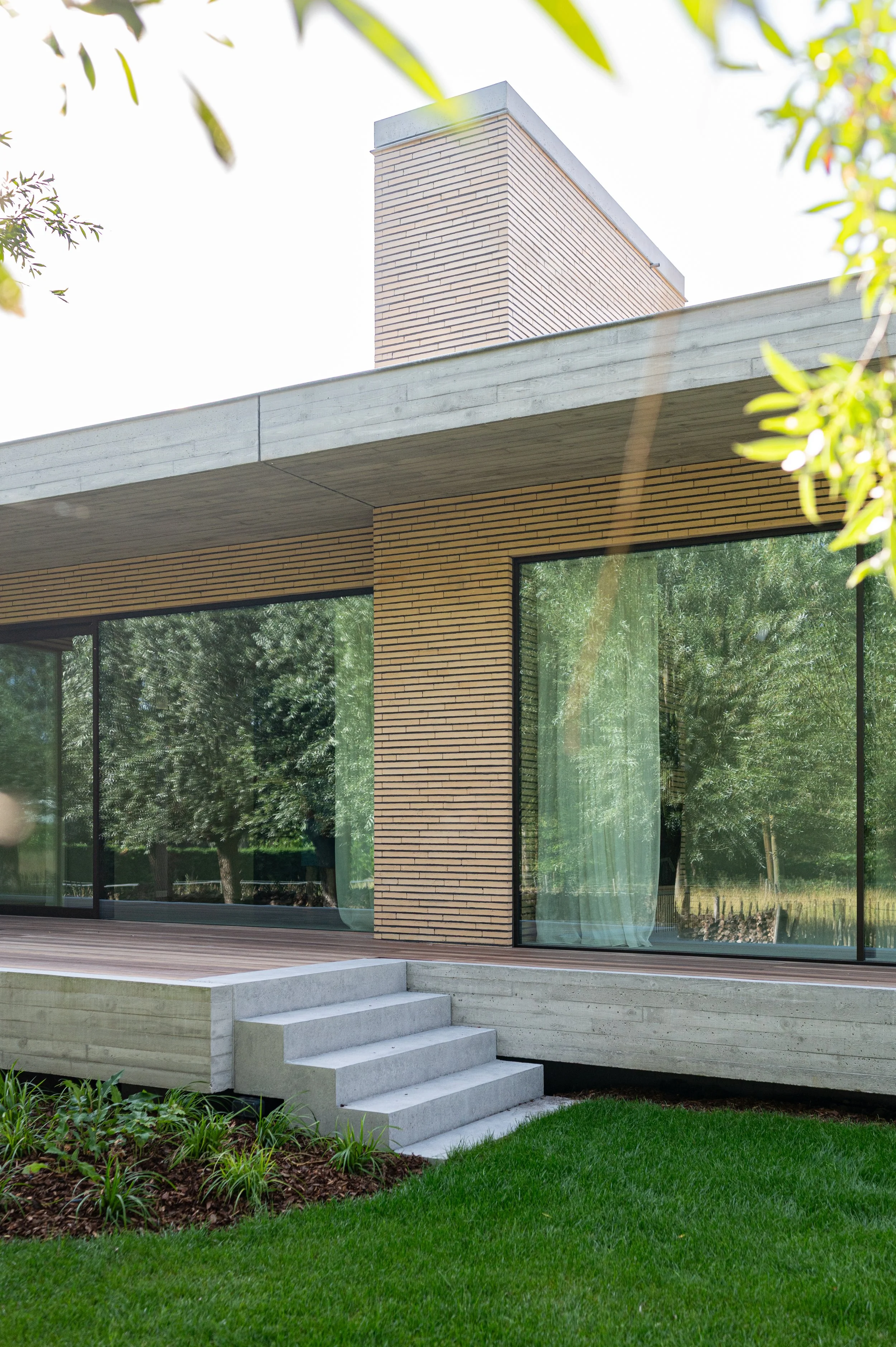 Modern house with brick and glass exterior, concrete steps leading up to a patio, large windows reflects trees, lush green lawn, and some branches with leaves in the foreground.