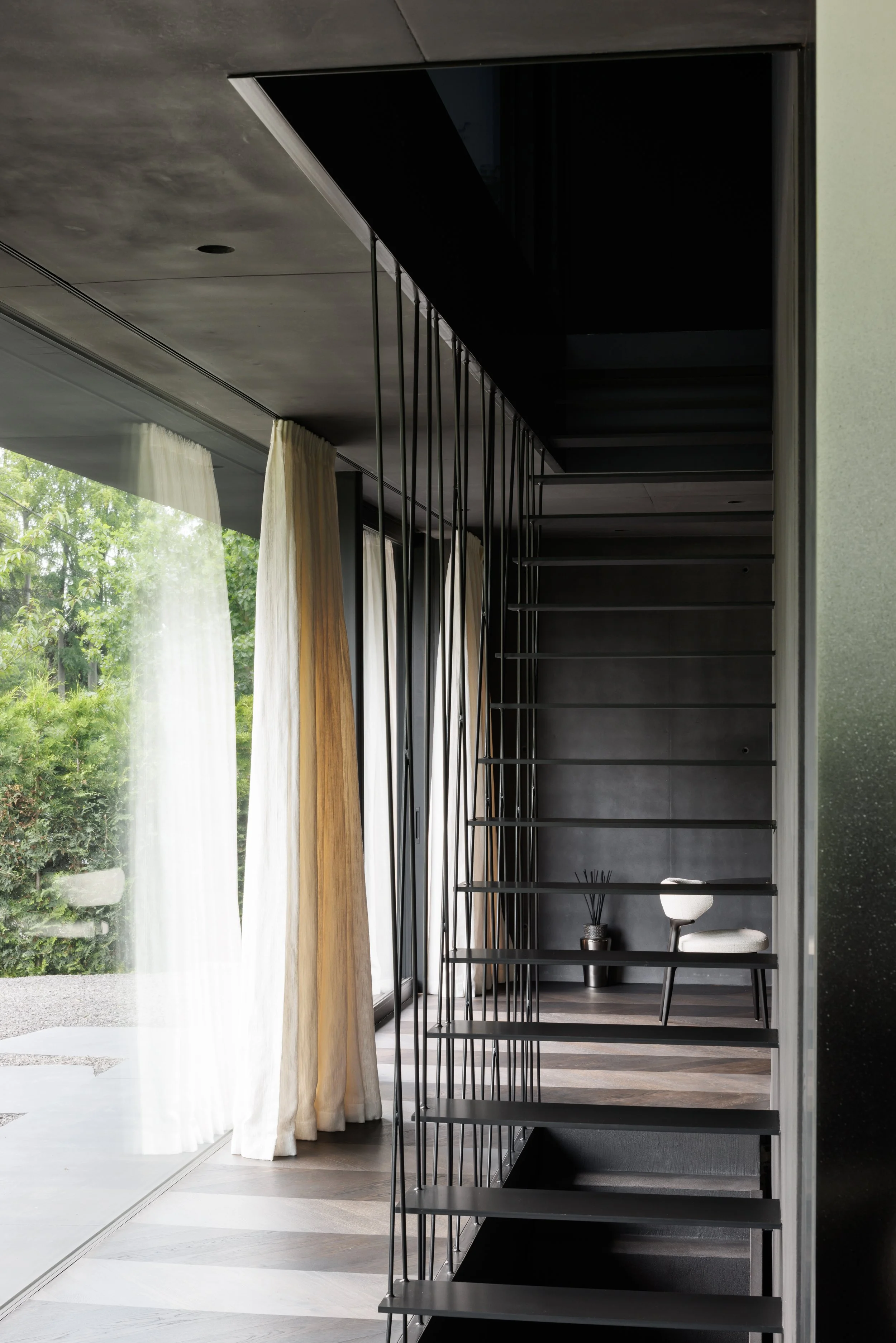 Modern interior staircase with black metal frame and wooden steps, next to a large window with white curtains, and a small sitting area with a chair and side table in a contemporary home.