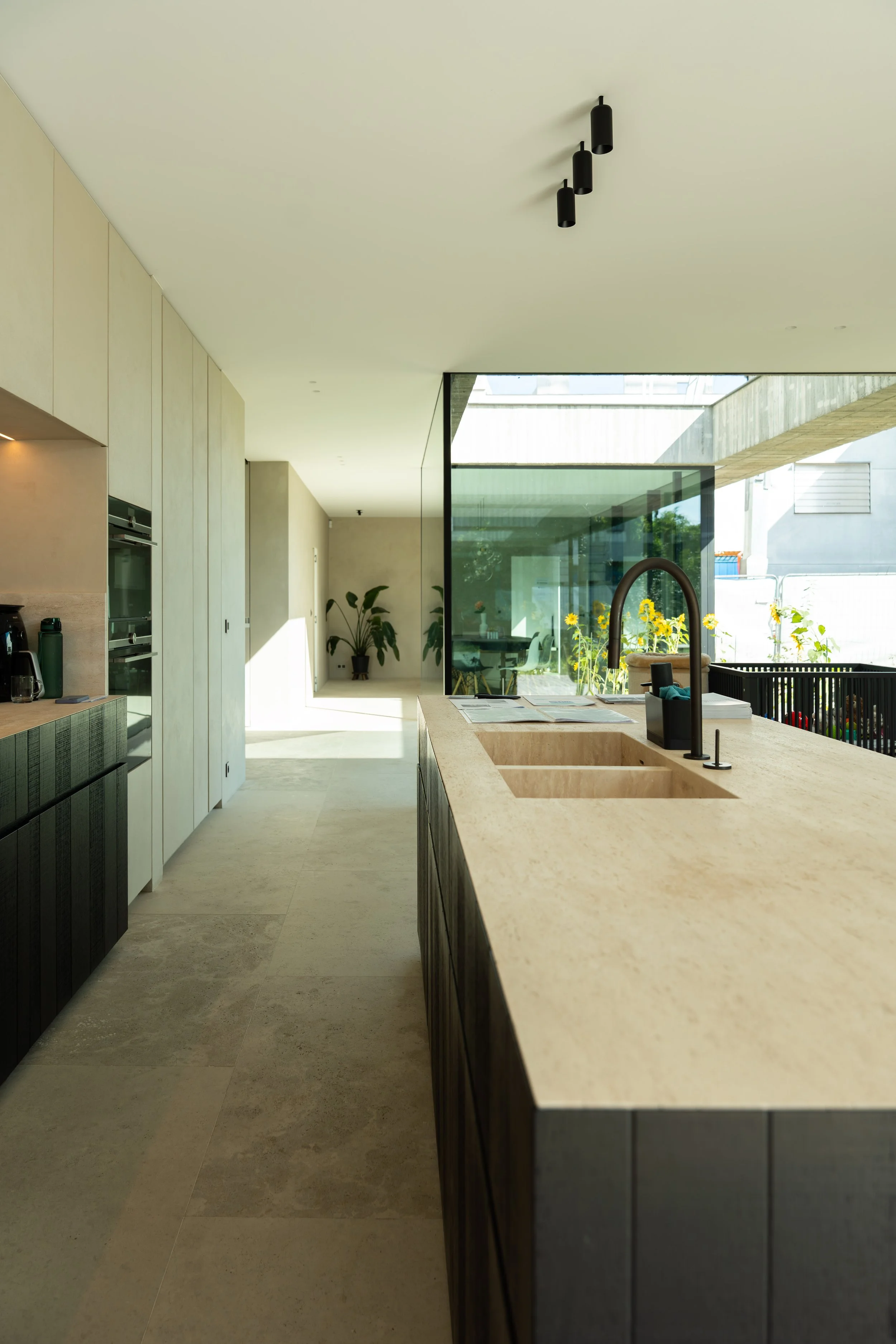 Modern kitchen with beige countertop island, black sink faucet, glass wall with sunflowers, and potted plants in the background.
