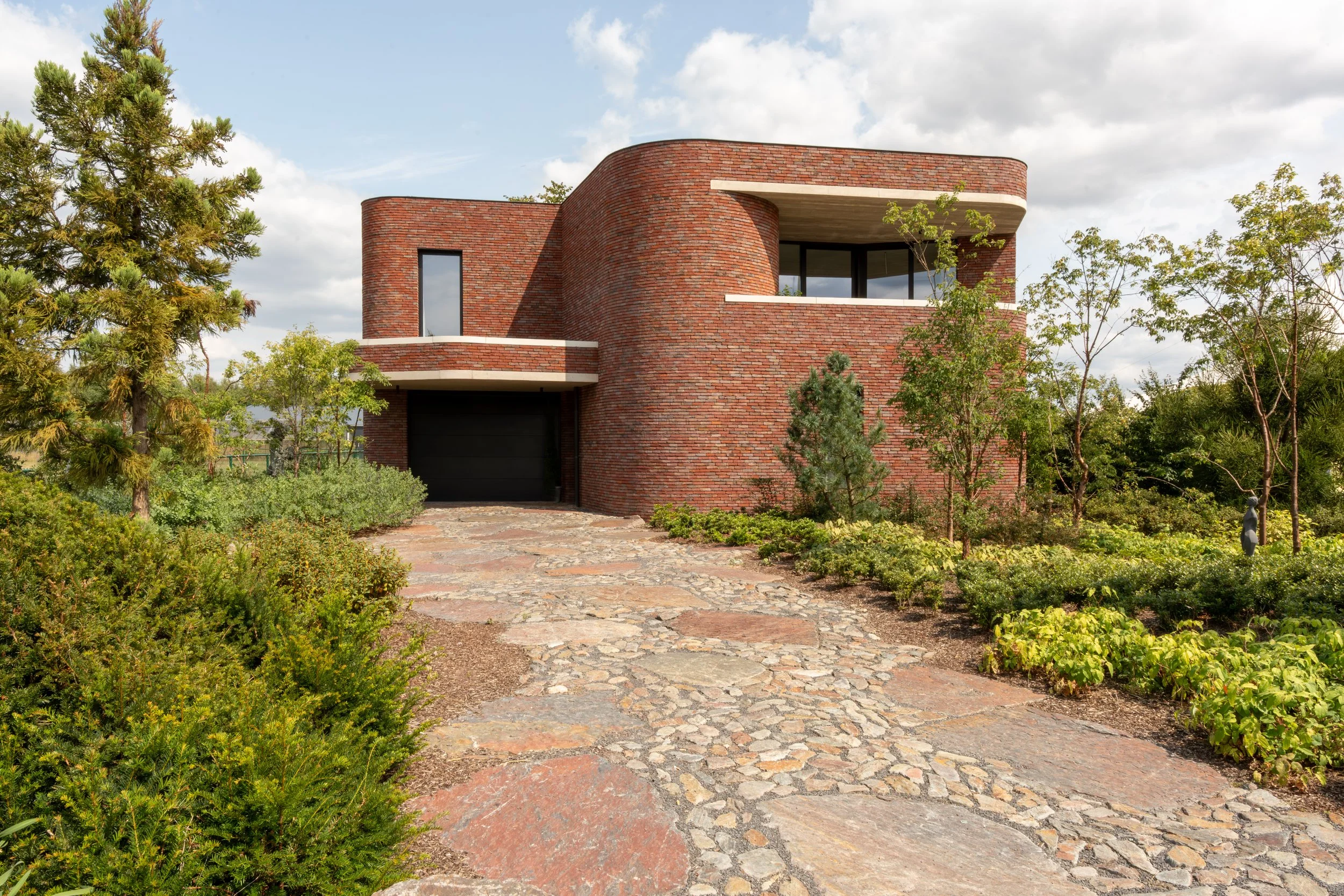 Modern red brick house with curved design, large windows, and a black garage door, surrounded by greenery and a stone pathway.