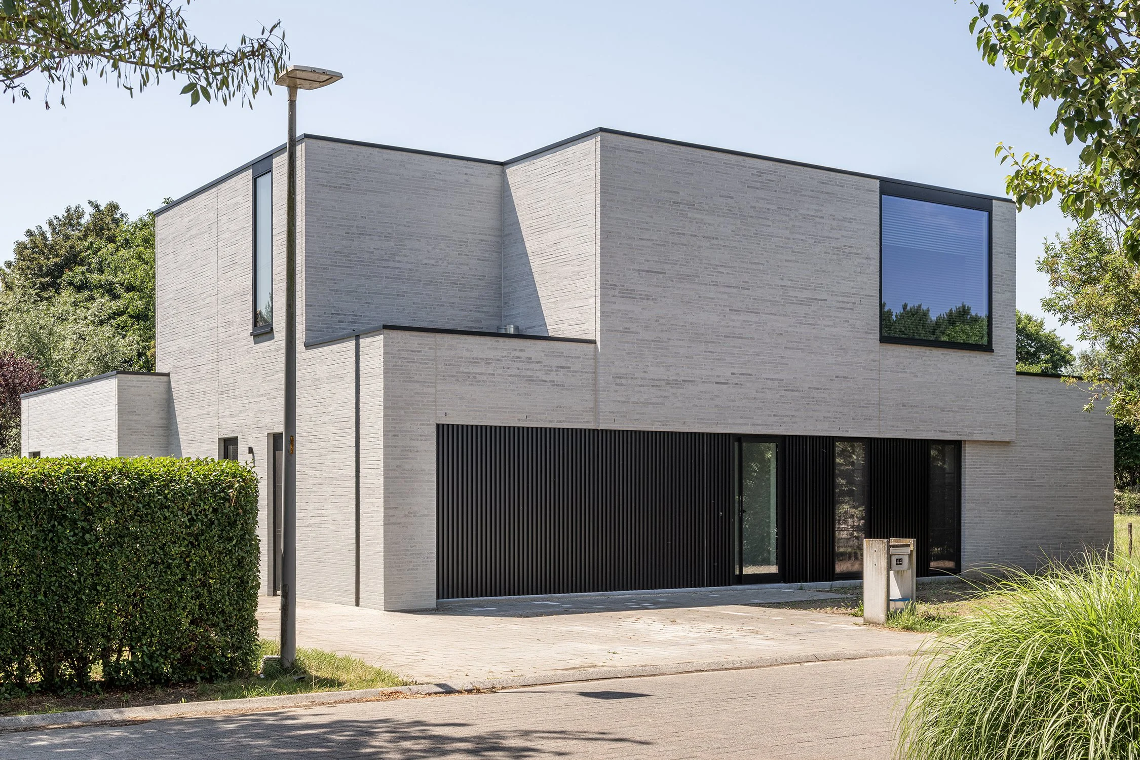 Modern two-story house with white brick exterior, large black-framed windows, black garage door, surrounded by greenery, with street and mailbox in foreground.