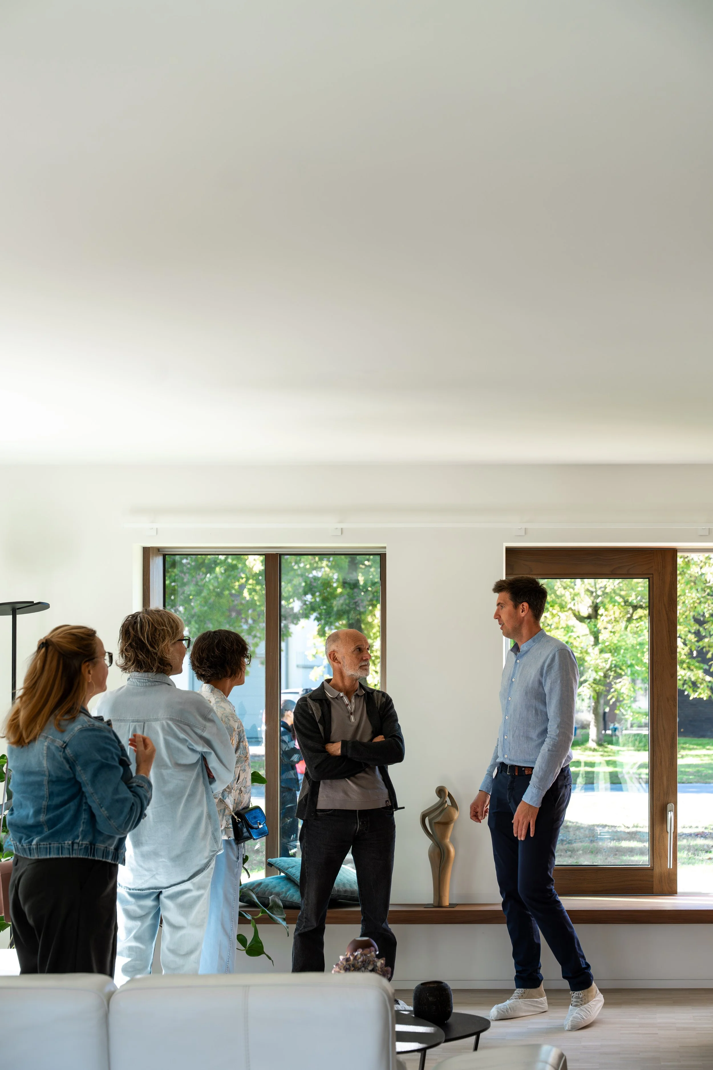 A man explaining something to a group of four women inside a modern living room with large windows and greenery outside.