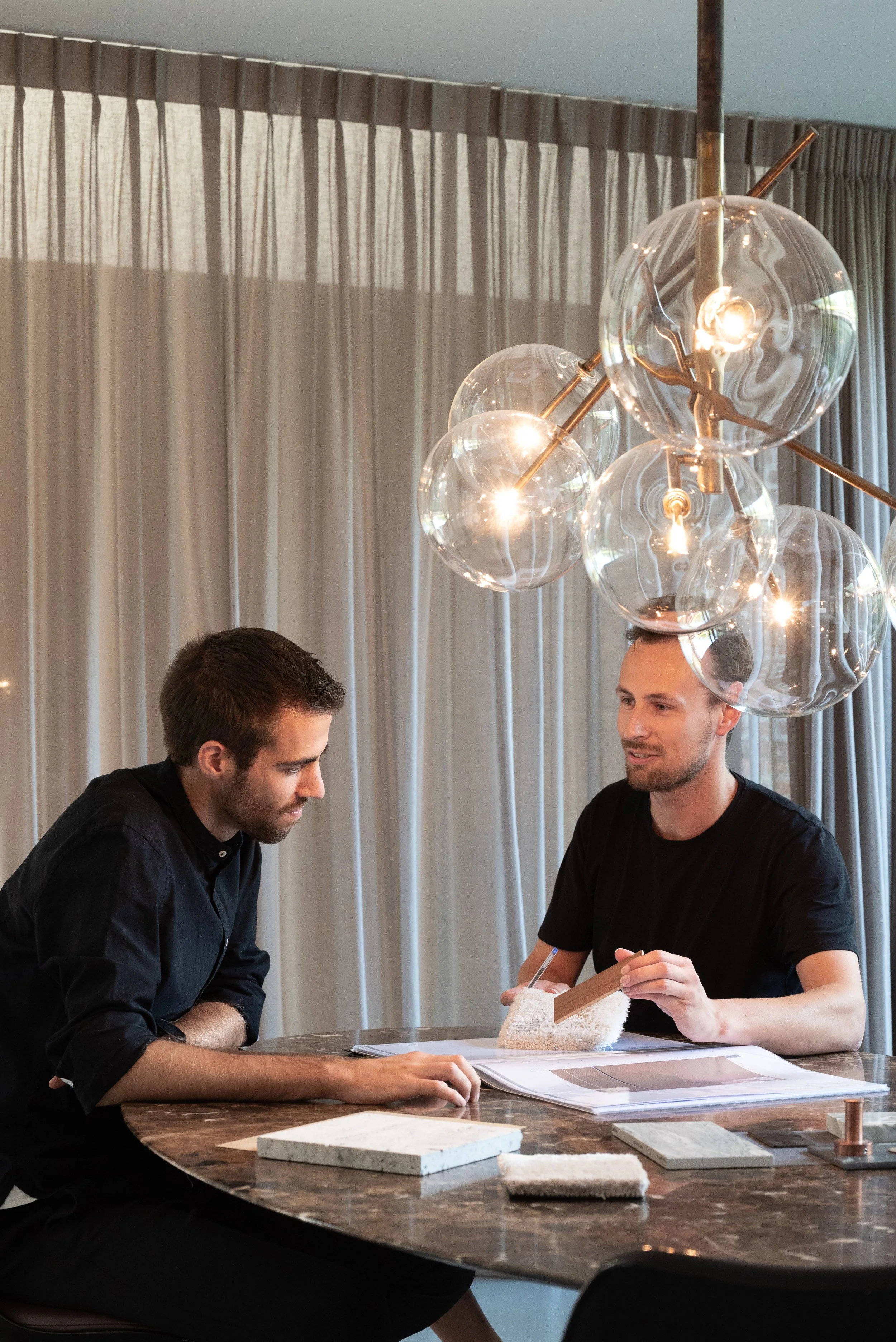 Two men are seated at a marble table discussing design samples, with materials and fabric swatches laid out, under a modern chandelier with glass globes and filament bulbs.