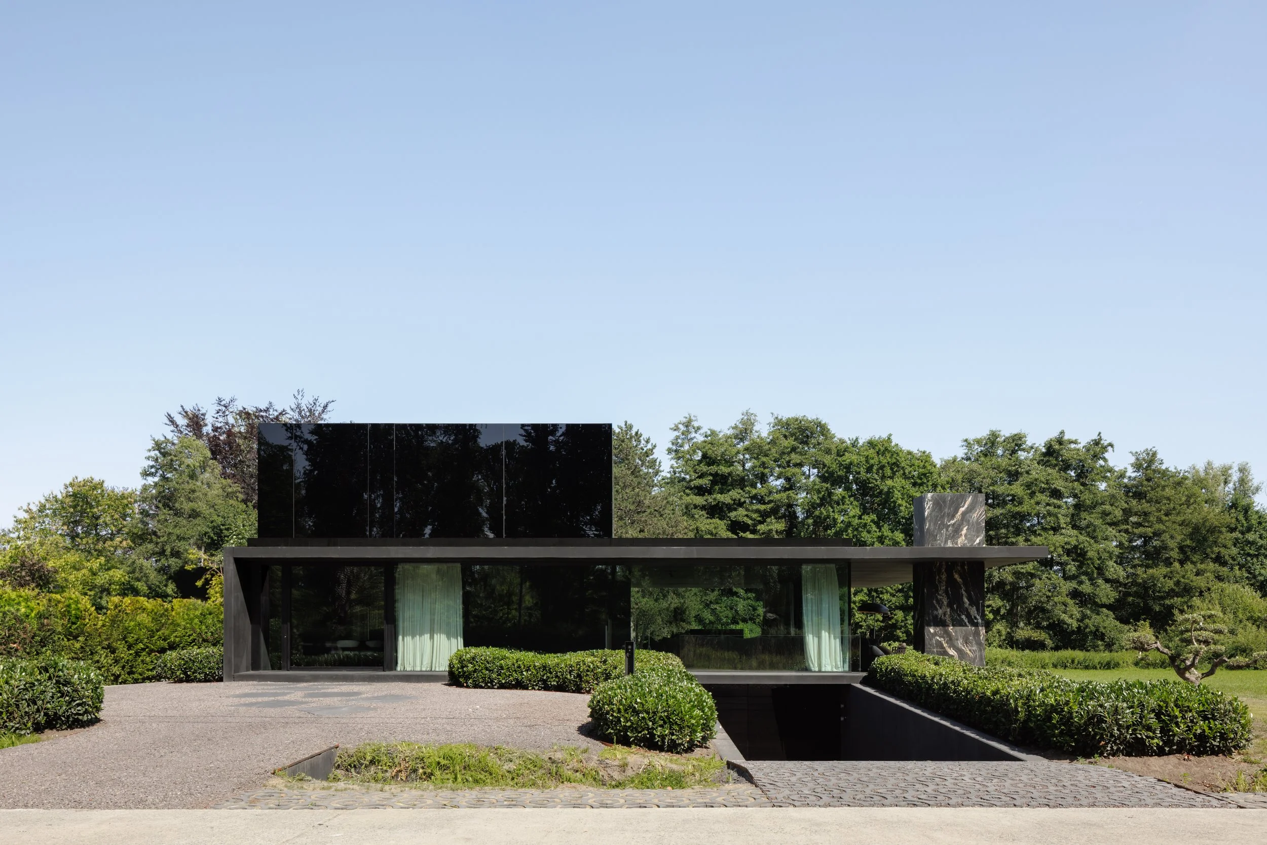 Modern black building with large glass windows, surrounded by green bushes and trees under a clear blue sky.