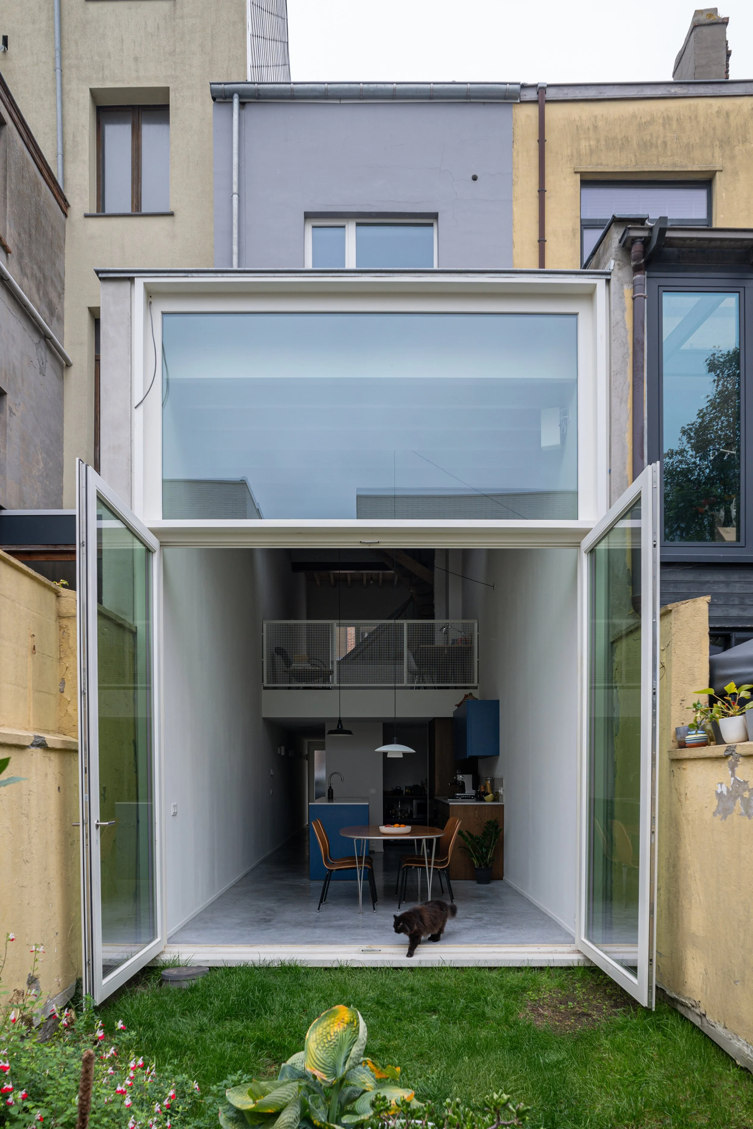 Open glass doors leading into a modern, minimalist kitchen and dining area with a small round table and three chairs, overlooking a small green backyard with a black cat walking across the floor.