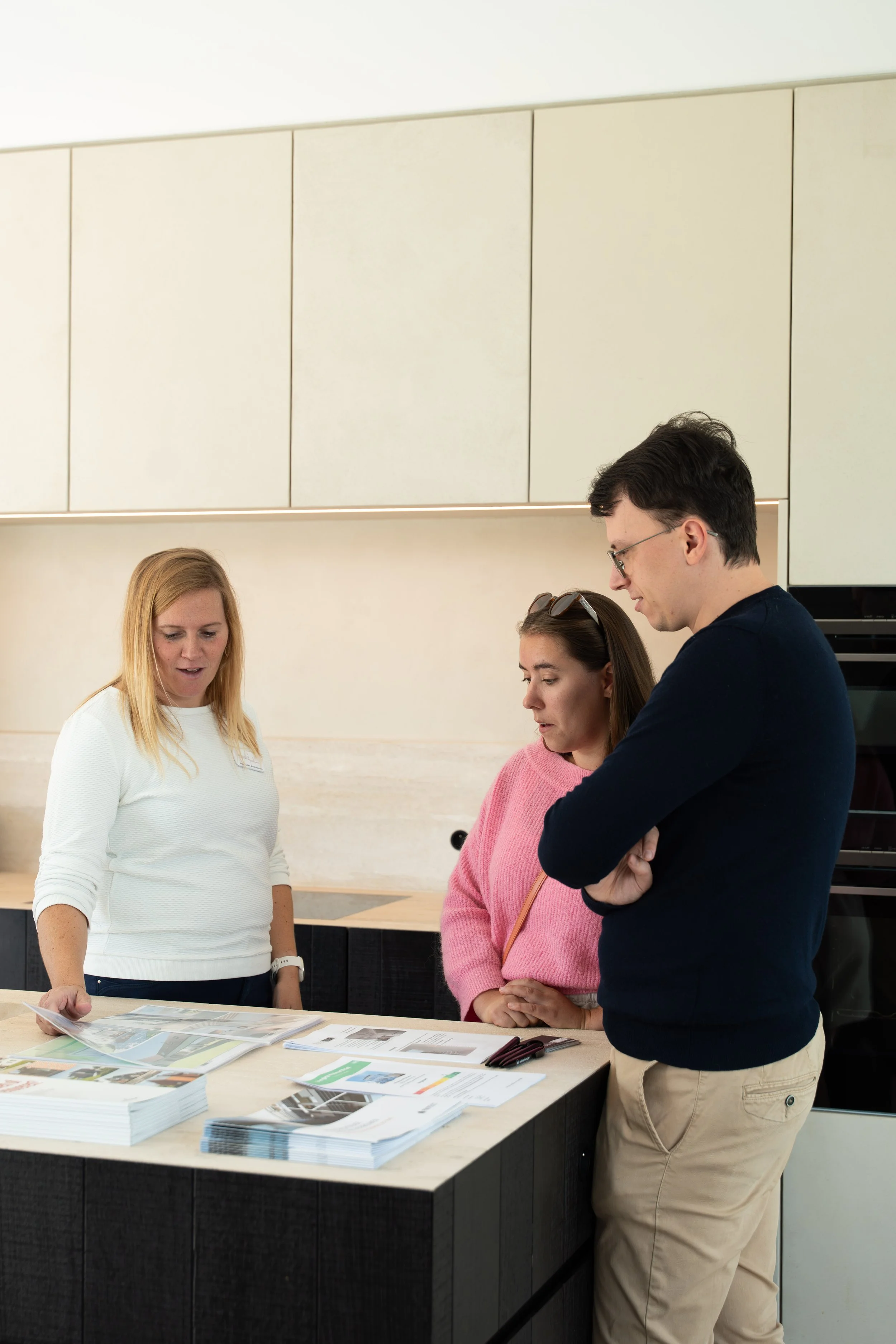 Three people looking at brochures and papers on a kitchen counter, engaged in discussion.