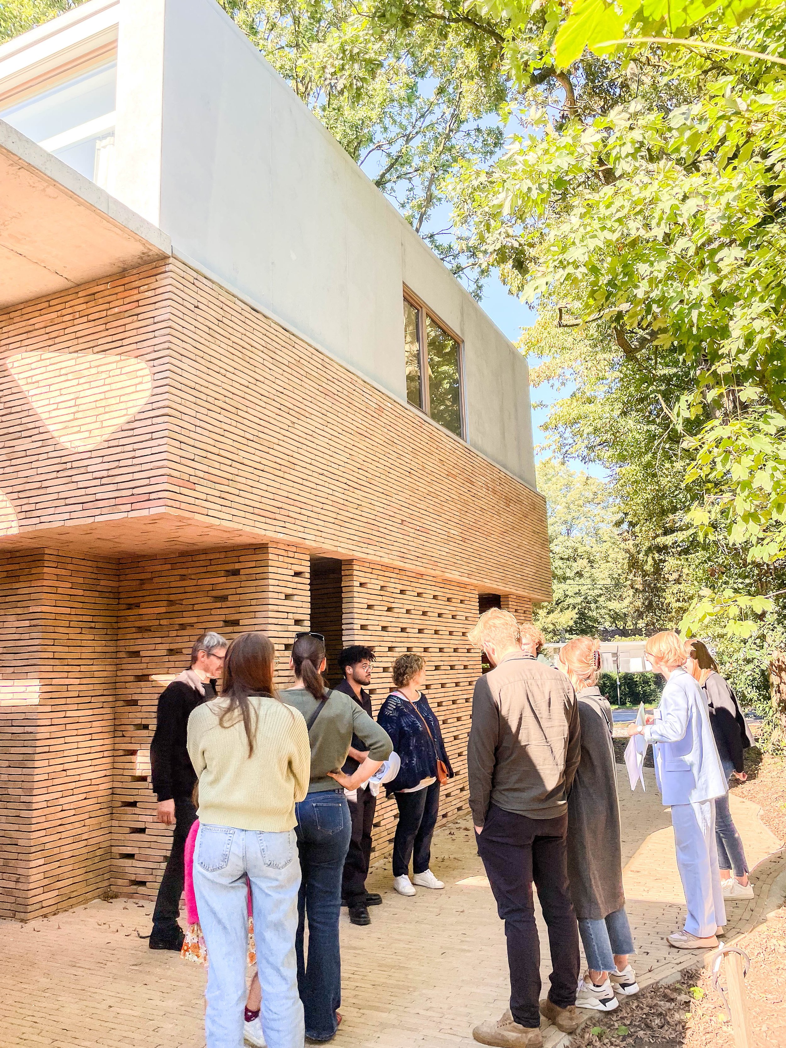 Group of people standing outside a modern brick building with large windows, some holding papers, under trees with green leaves.