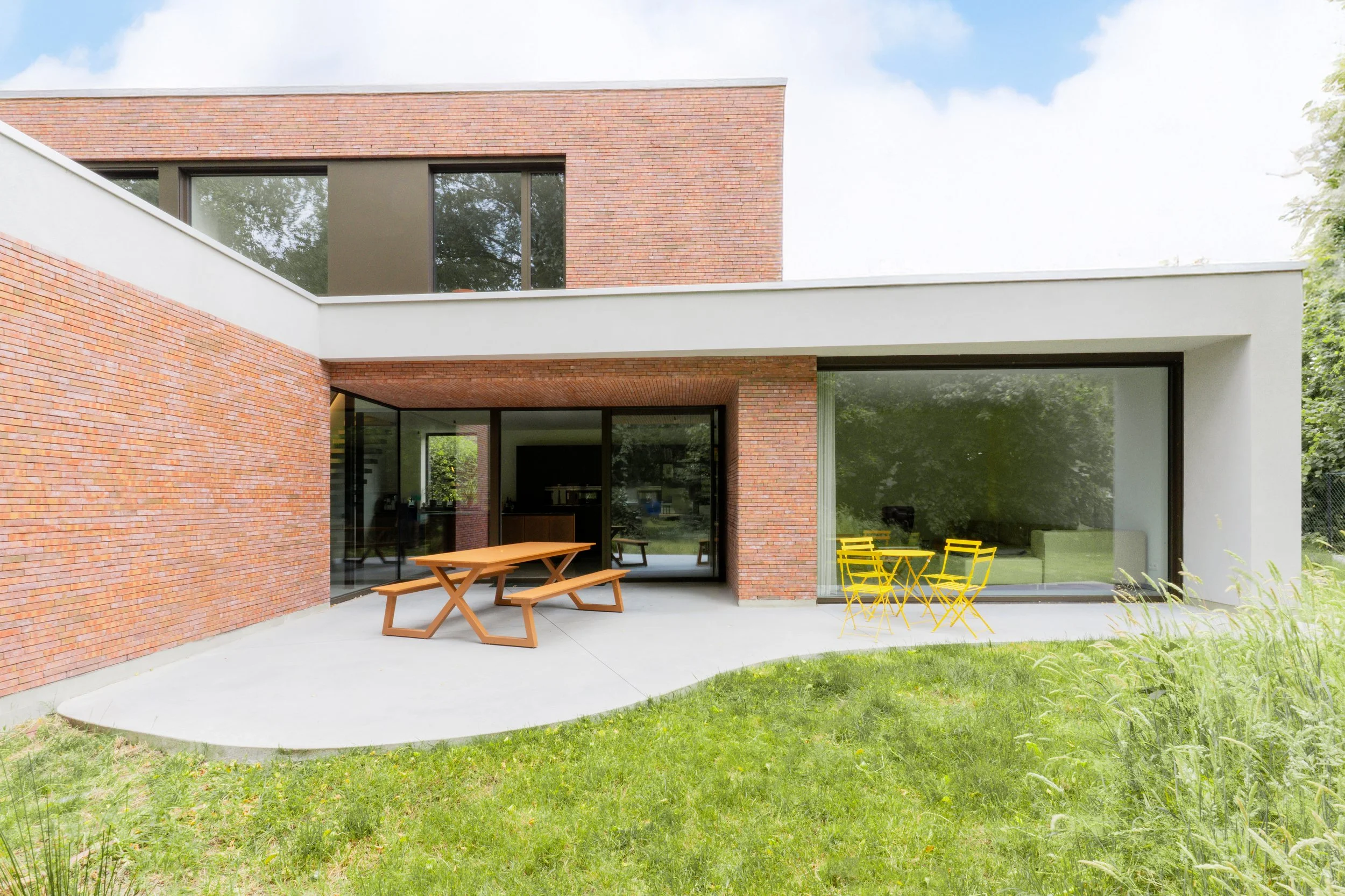Modern house with large glass sliding doors, brick walls, and a grassy backyard featuring a wooden picnic table and yellow metal outdoor furniture.