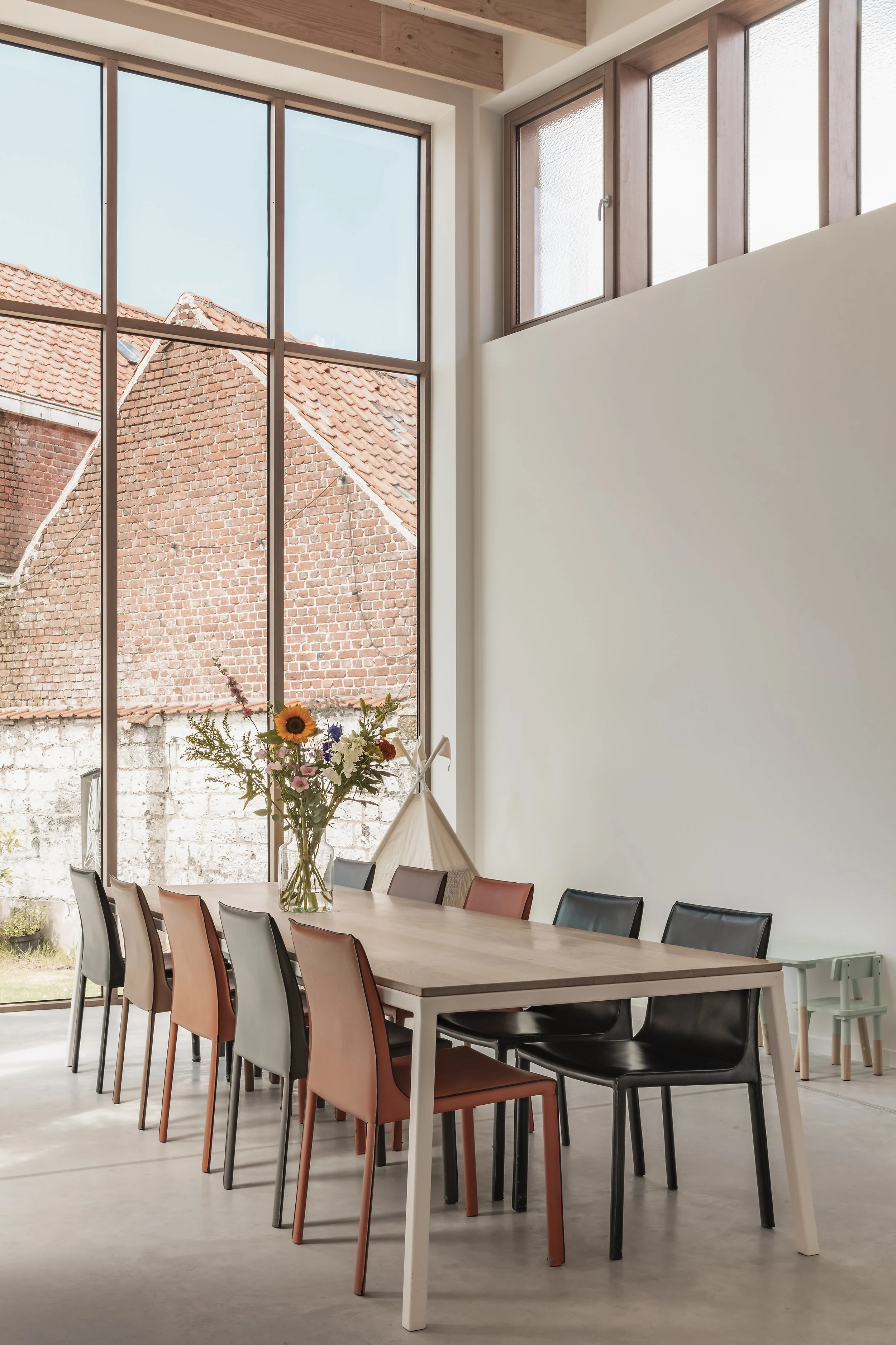 A dining room with a long wooden table, colorful chairs, a large window, a vase of sunflowers and flowers, and a fabric teepee in the corner.
