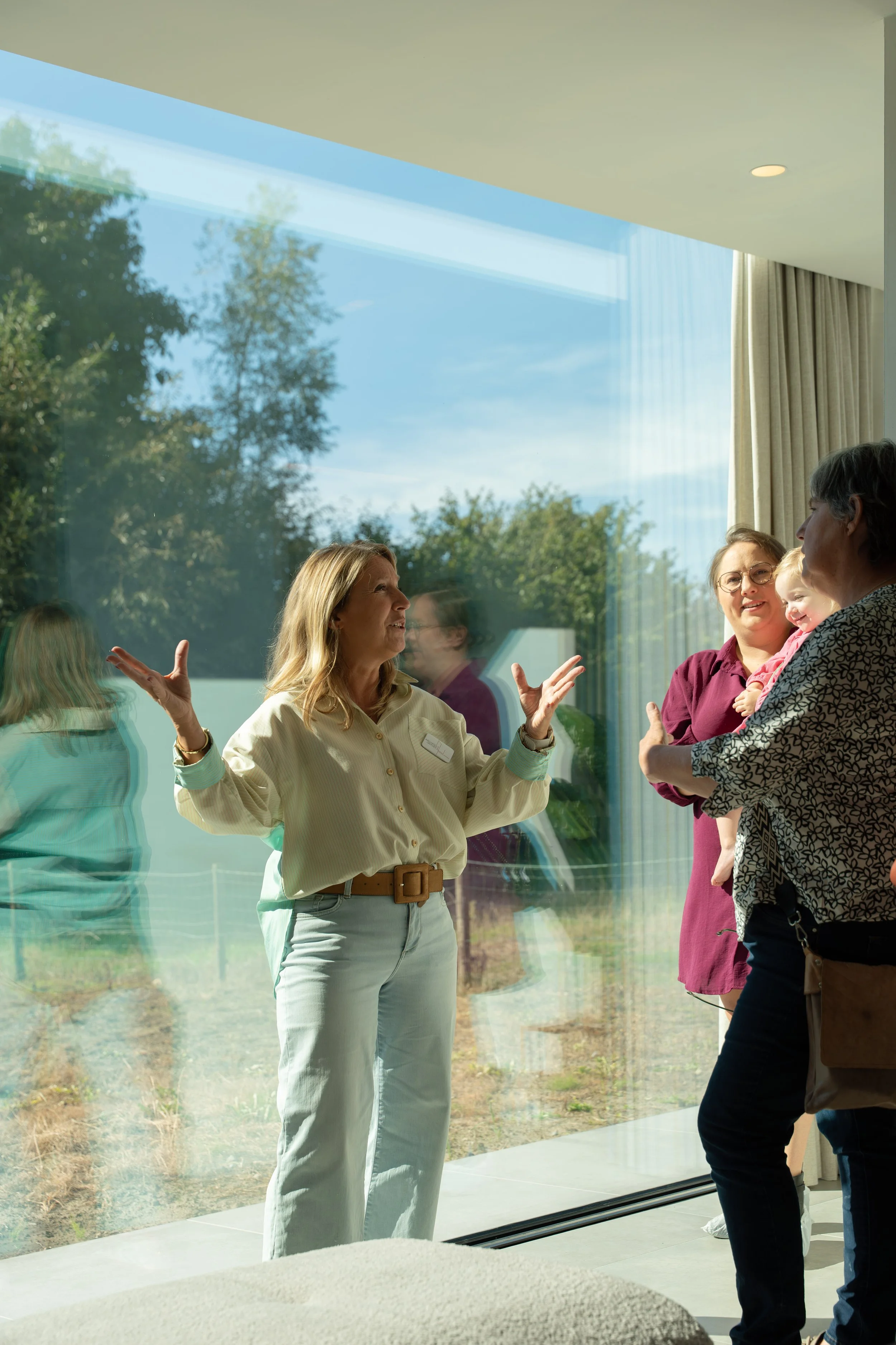 Two women are engaged in conversation through a large glass window, with a third woman and a child inside the room. The scene is well-lit with natural light, and trees are visible outside.