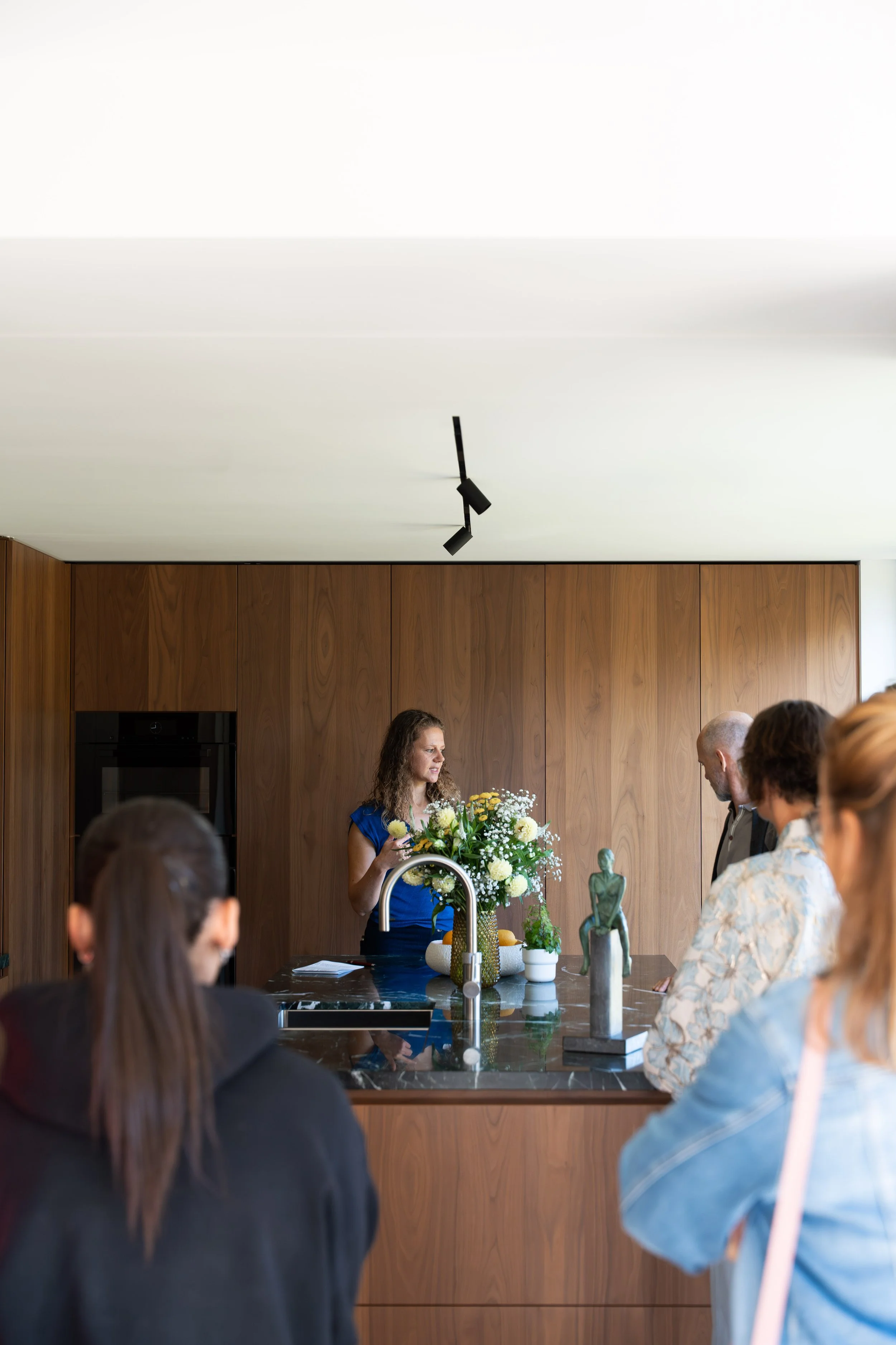 A woman with curly hair giving a presentation in a modern kitchen with wooden cabinets, a marble countertop, and a large bouquet of flowers. Several people are listening, and artwork and decorative sculptures are on the counter.