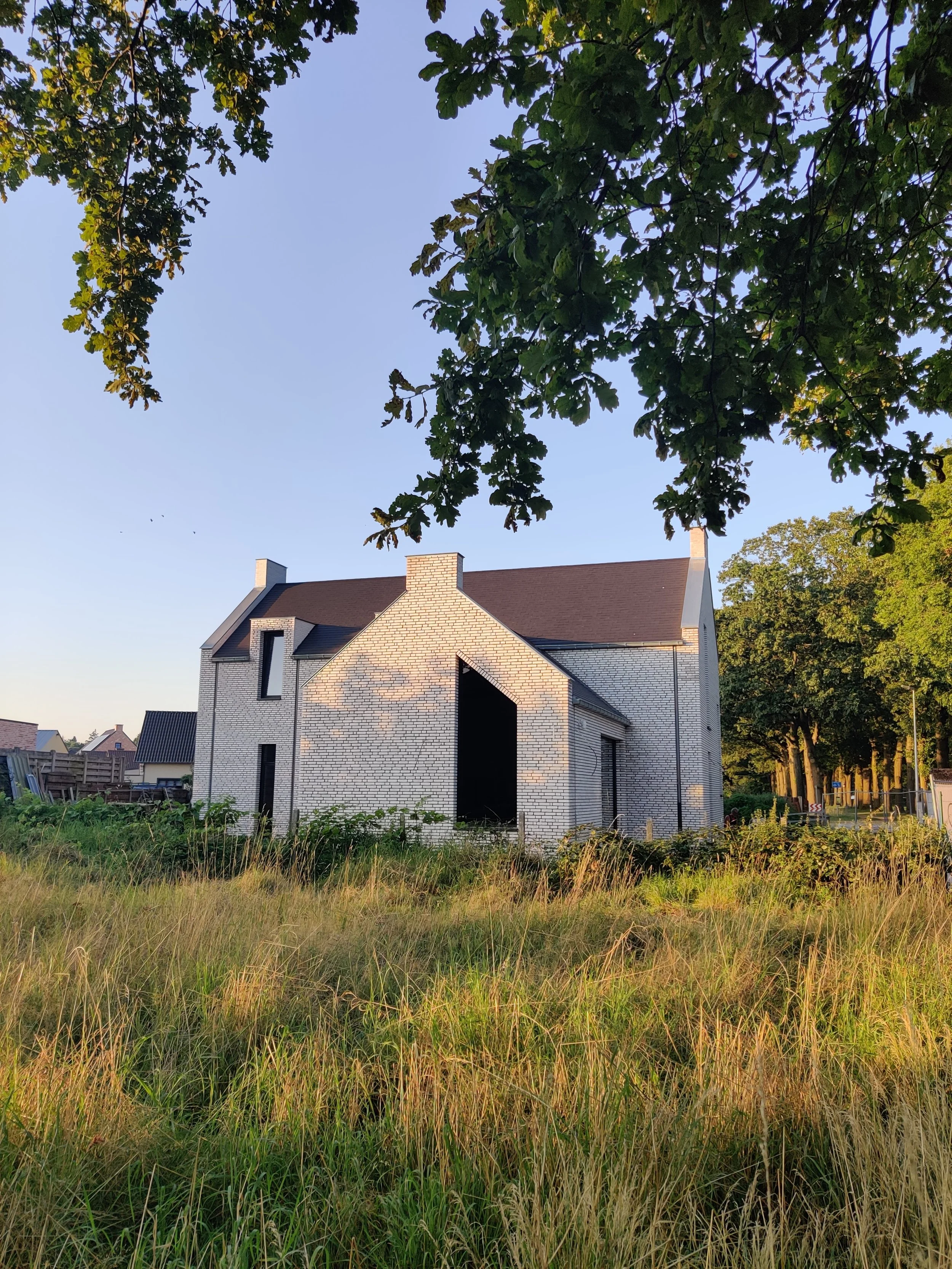 A house under construction with white brick walls and a brown roof, surrounded by tall grass and trees, with a clear blue sky above.