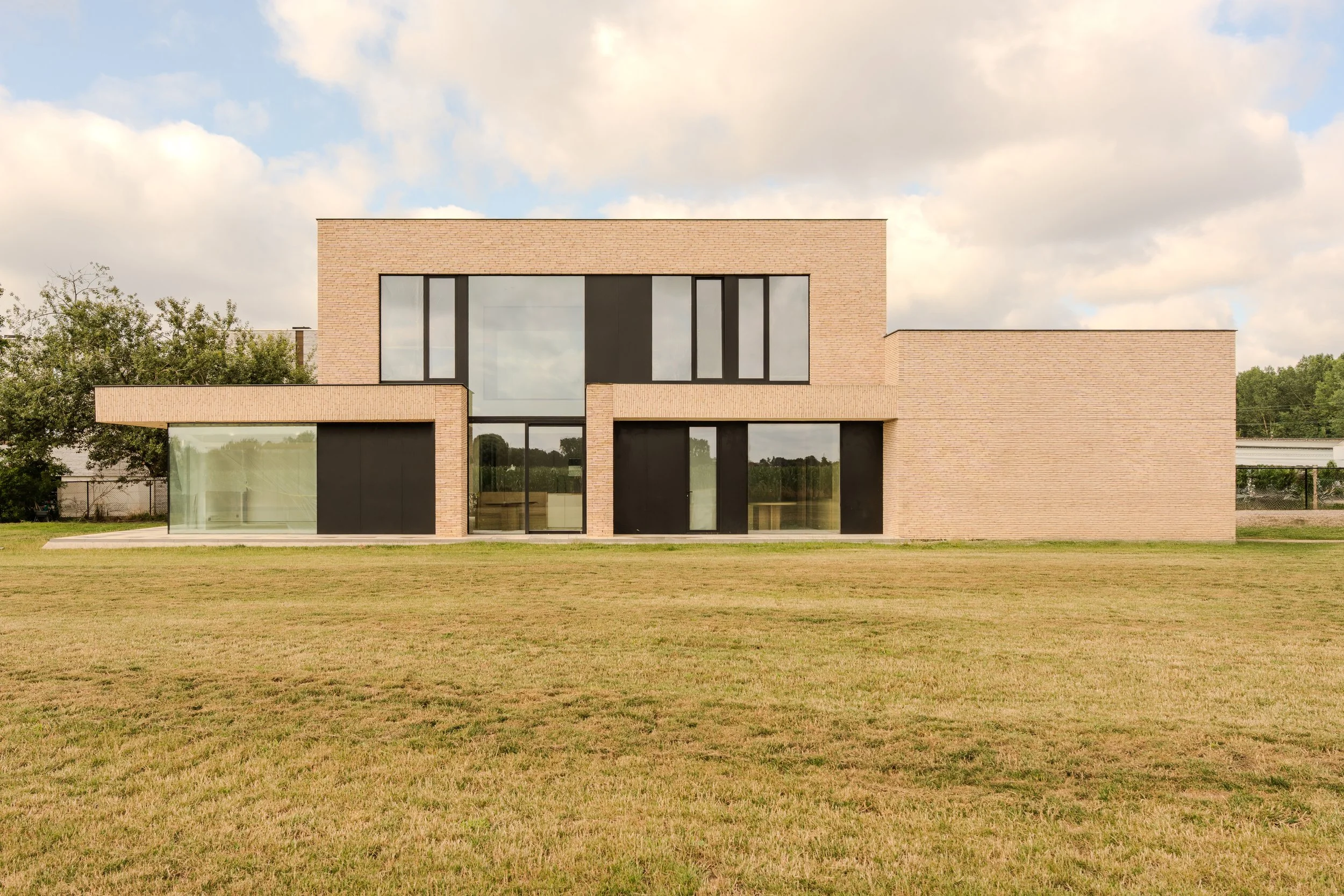 A modern, two-story brick house with large black window frames, situated on a grassy lawn under a partly cloudy sky.