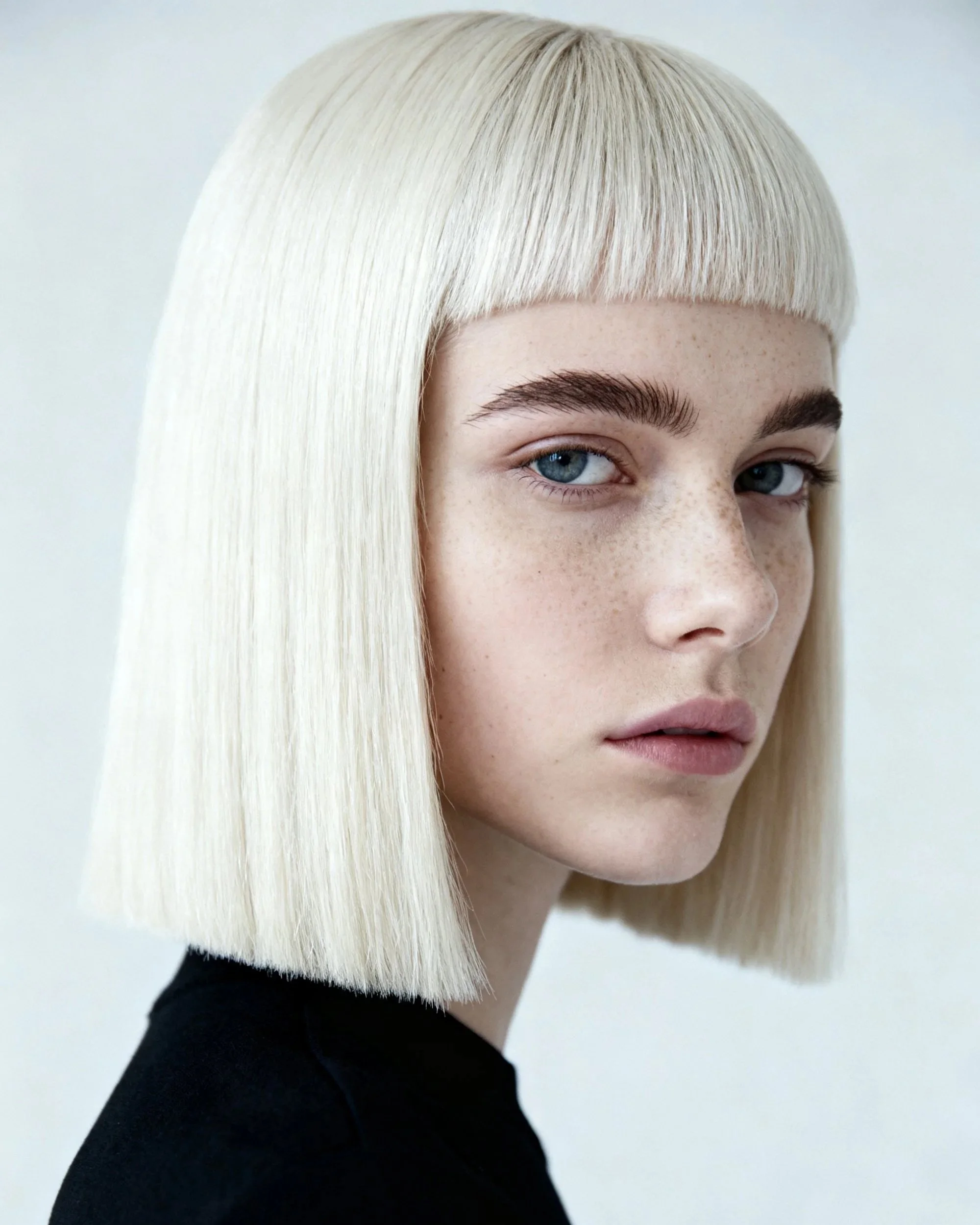 Close-up of a woman with platinum blonde hair, styled in a sleek, straight bob with blunt bangs, wearing a black top against a plain light background.