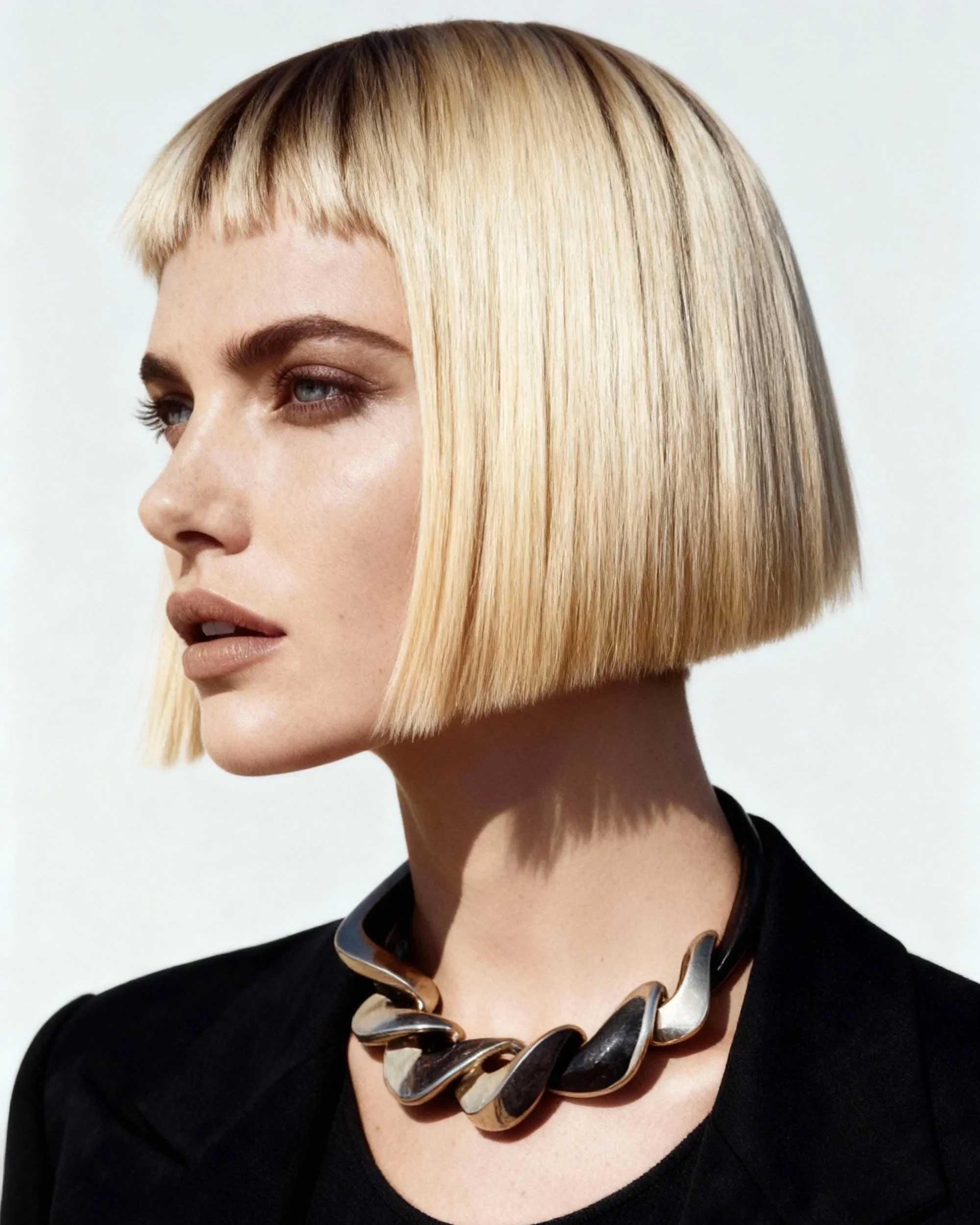 Close-up portrait of a woman with a short blonde bob hairstyle, wearing a black top and a chunky, black and silver statement necklace.