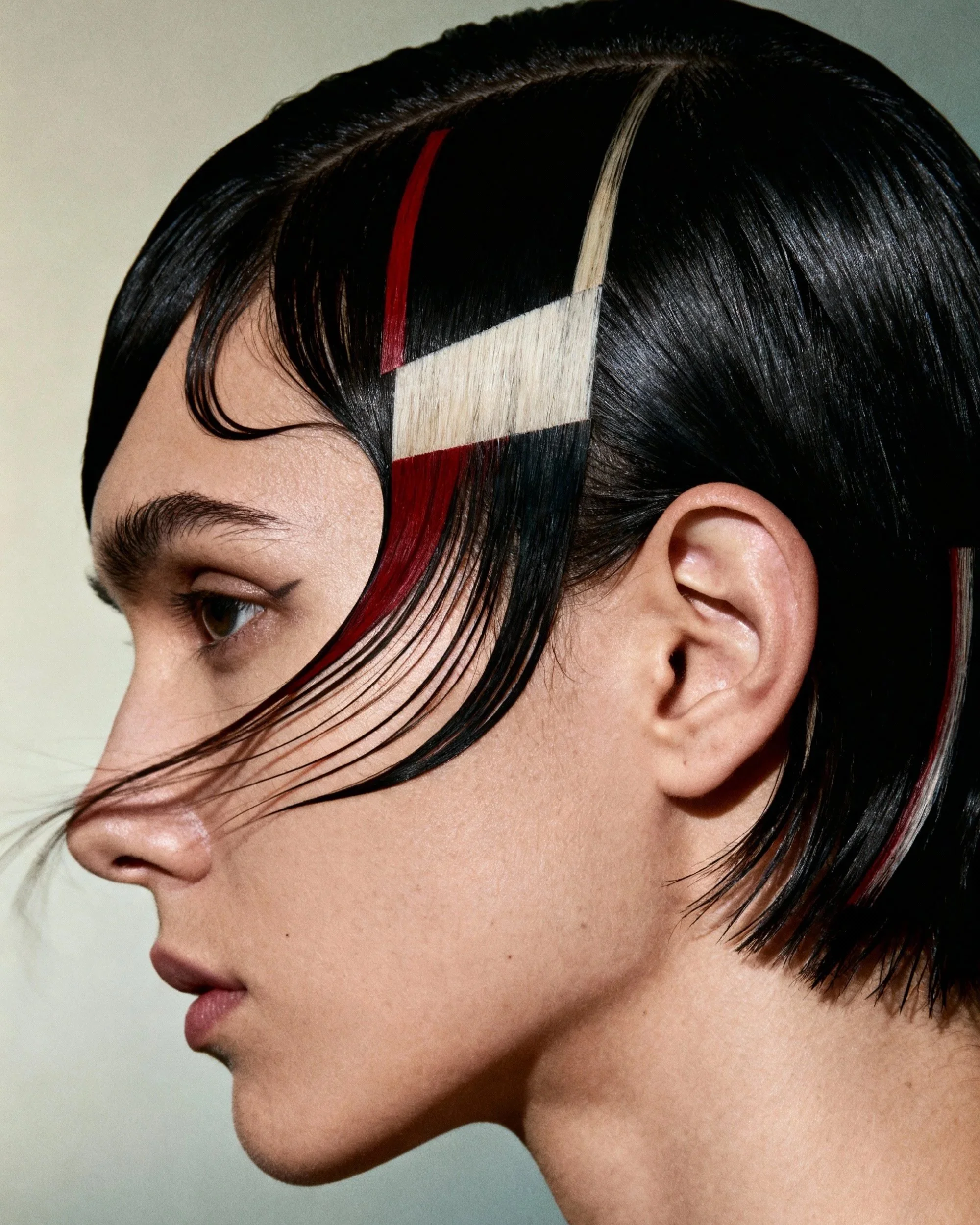 Close-up side profile of a woman with black hair with streaks of blond and red, styled sleek and straight, against a plain background.