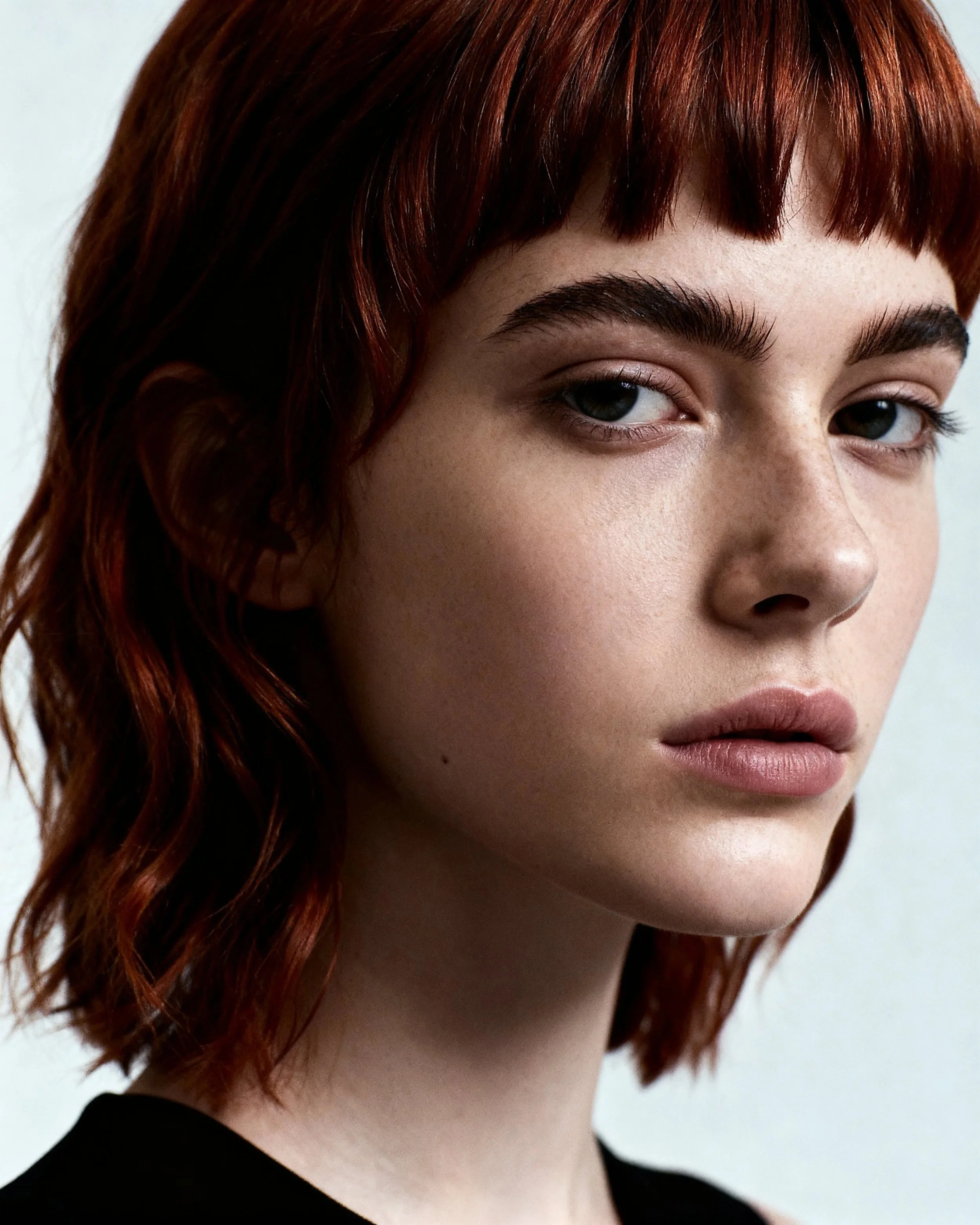 Close-up of a young woman with red hair, fair skin, and natural makeup, wearing a black top, against a plain light background.