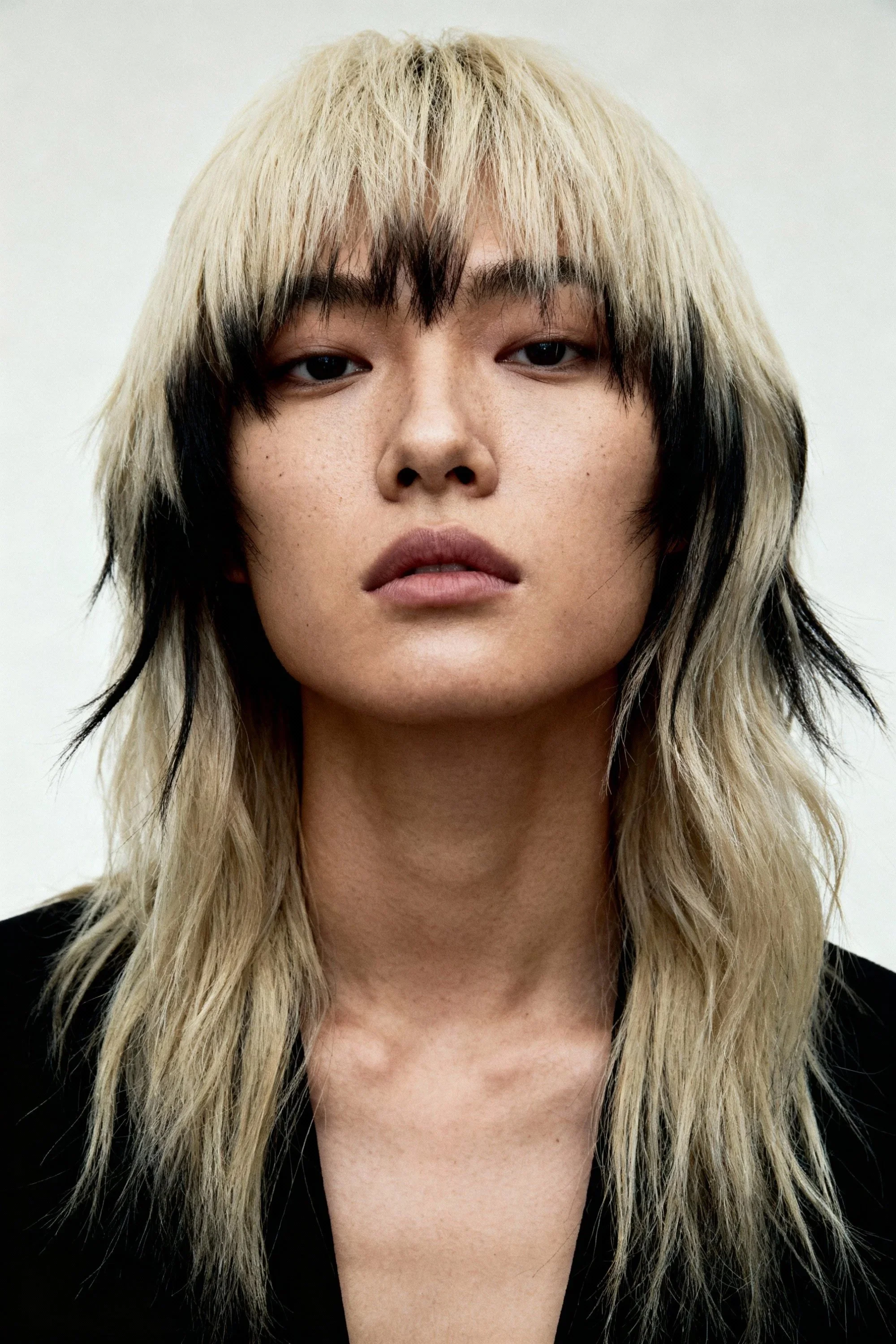 Close-up of a young person with long, layered blonde hair with dark roots, wearing a black top, and having a neutral facial expression, against a plain white background.