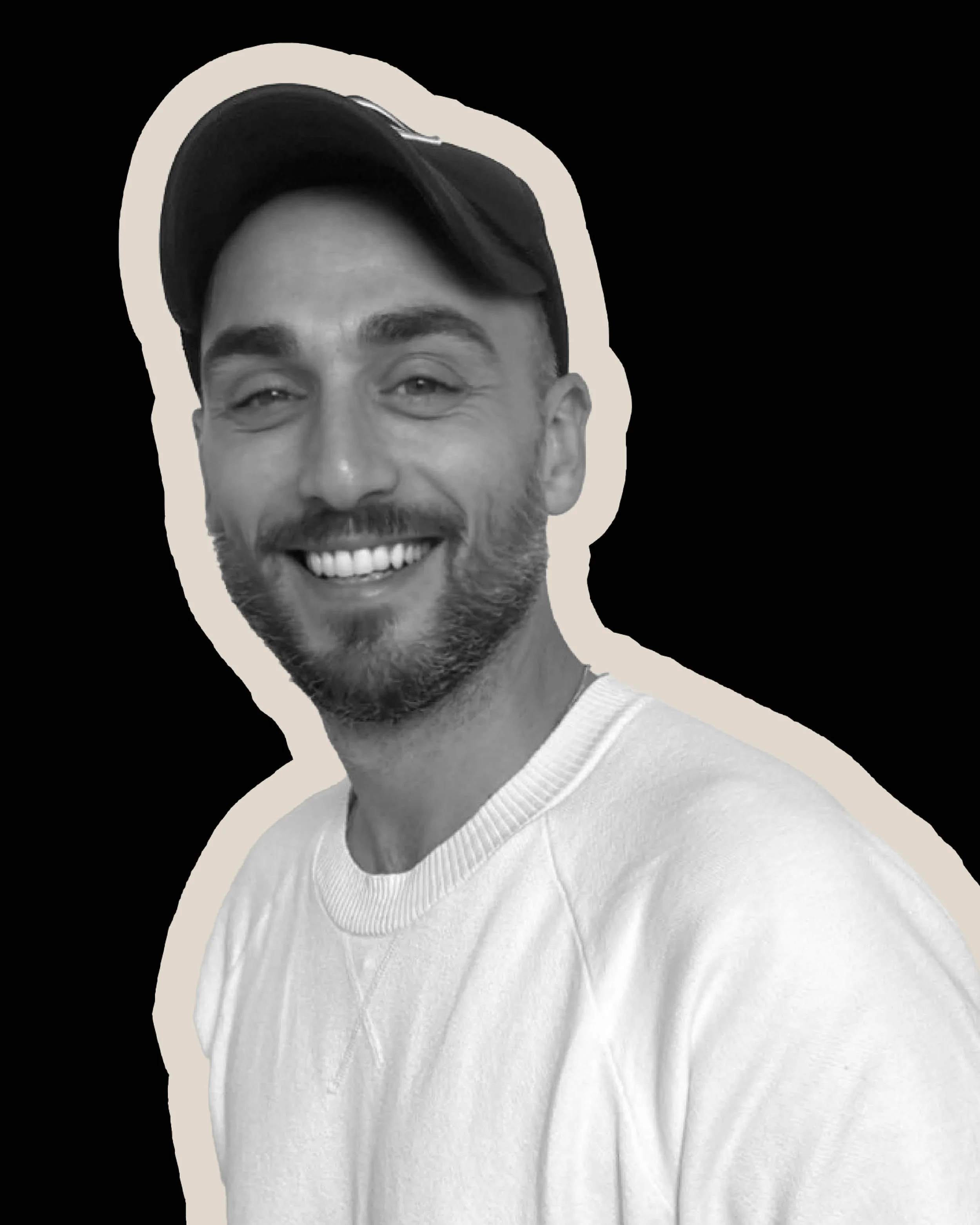 Black and white photo of a smiling man with a beard wearing a cap and a white sweater, with a beige outline around the image.
