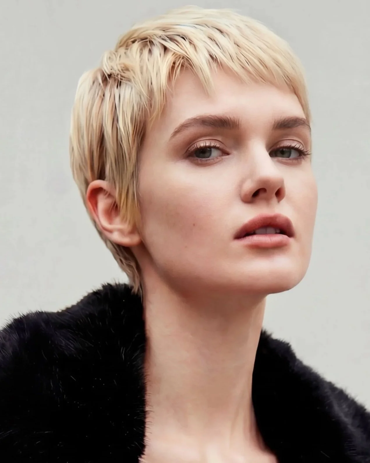 Close-up of a woman with short blonde hair, minimal makeup, wearing a black fur garment, against a plain light background.
