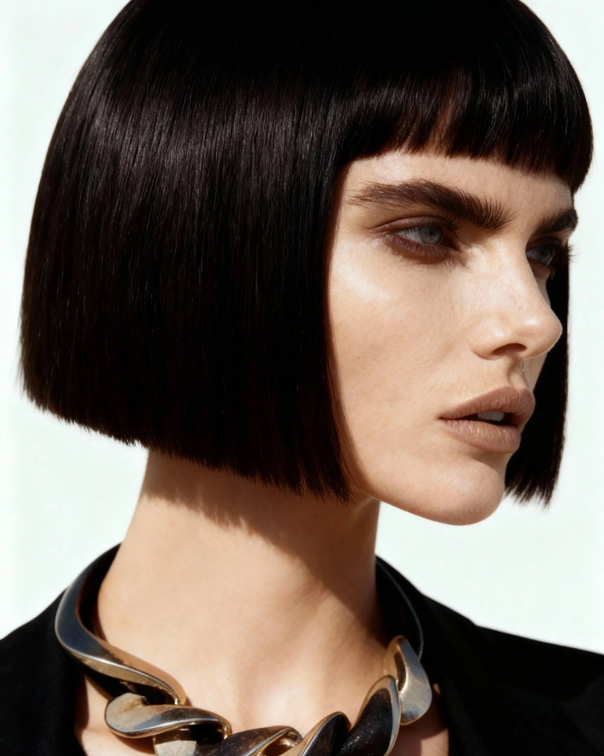 Close-up of a woman with straight dark hair and blunt bangs, wearing a black top and a chunky chain necklace.