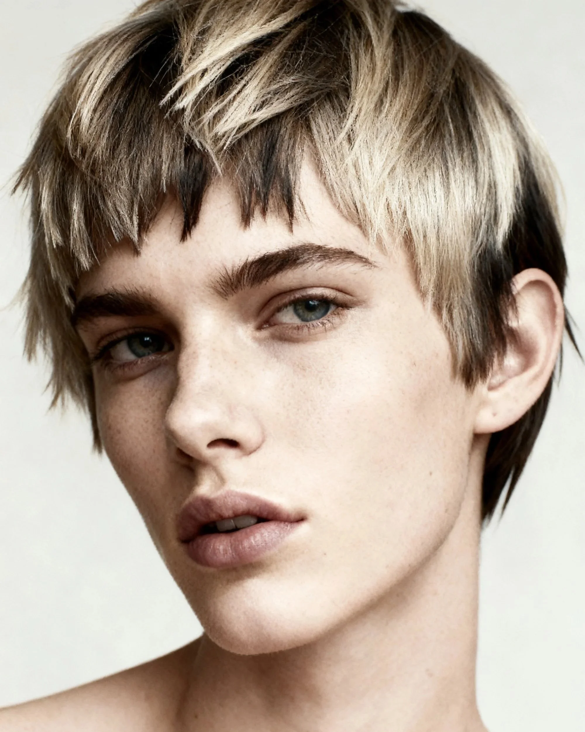 Close-up of a woman's face with short, layered, blonde and brunette hair, blue eyes, and pink lips, against a plain light background.
