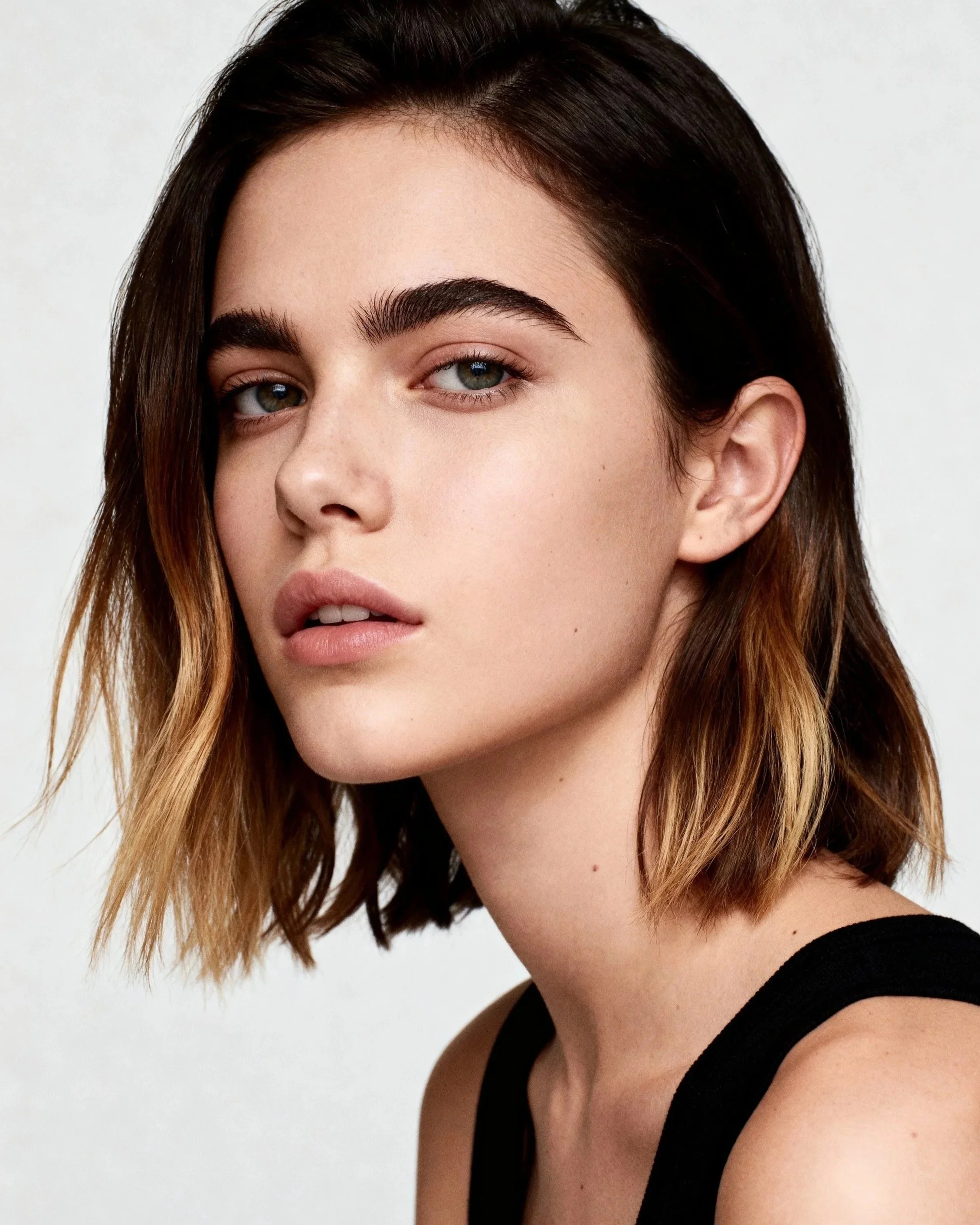 Close-up of a woman with short, wavy brown hair with blonde tips, wearing a black sleeveless top, posing against a white background.