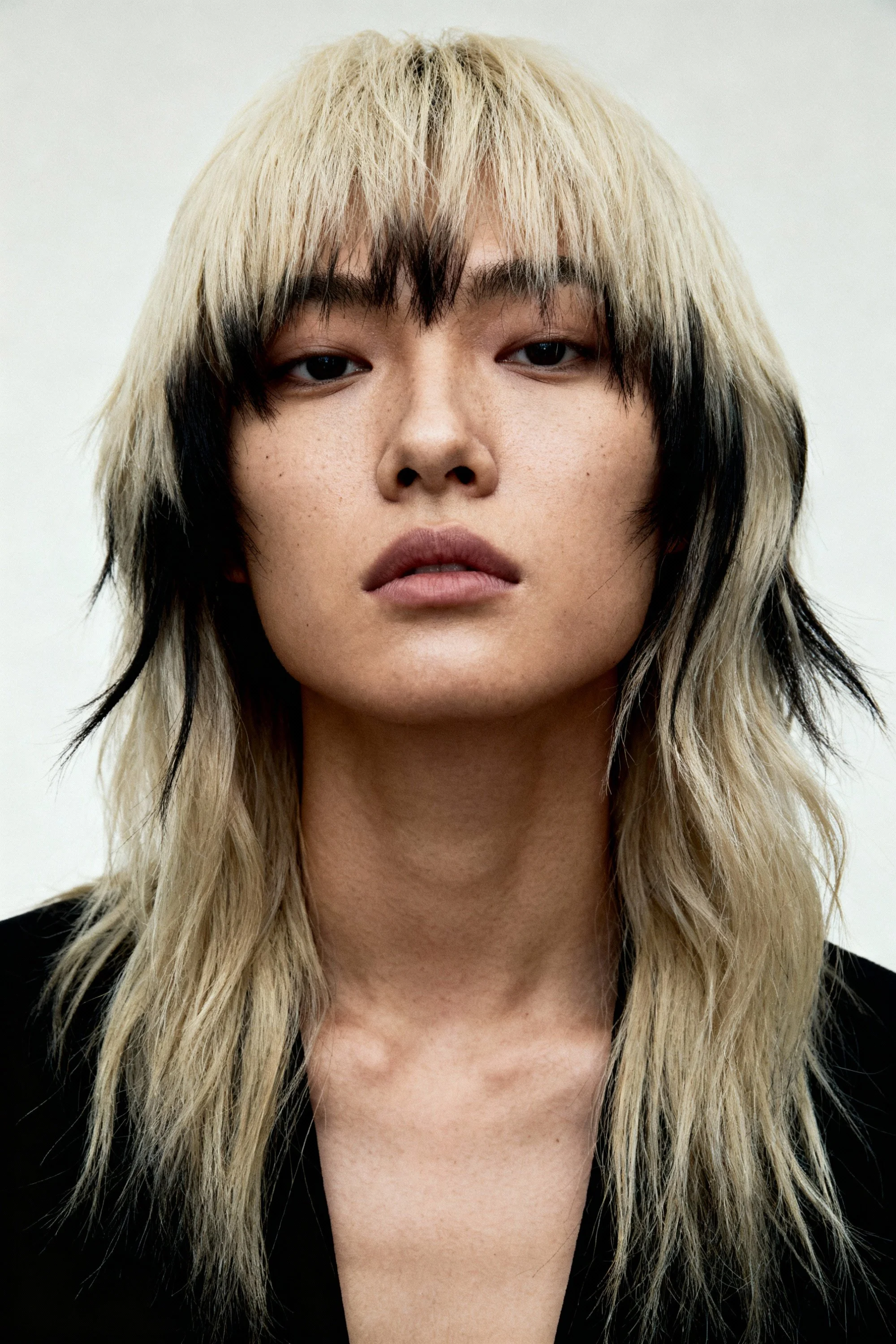 Close-up of a young person with long, layered blonde and black hair, wearing a black top, with a neutral facial expression