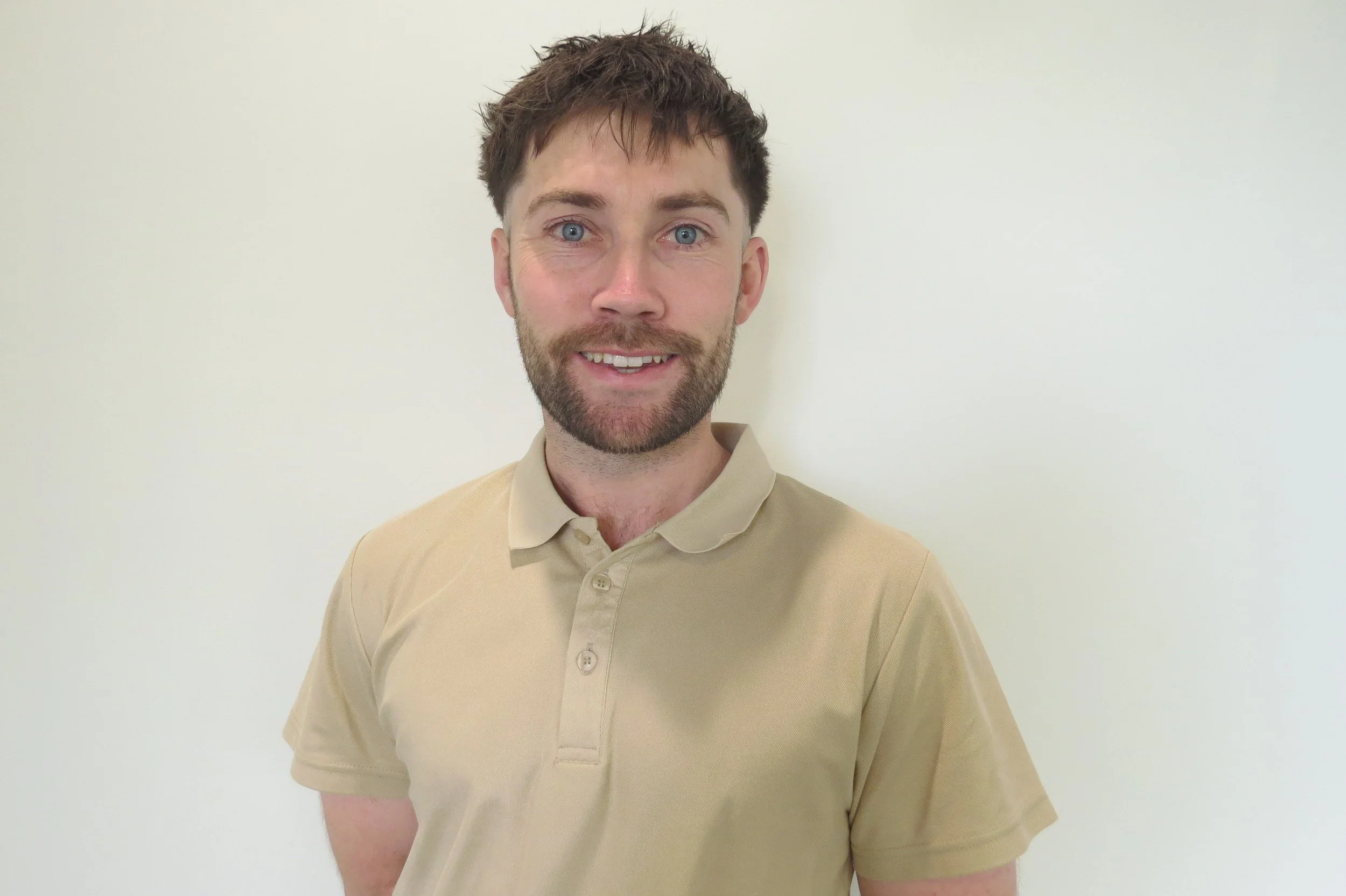 A young man with blue eyes, brown hair, and a beard, smiling, wearing a beige polo shirt, standing against a plain, light-colored wall.
