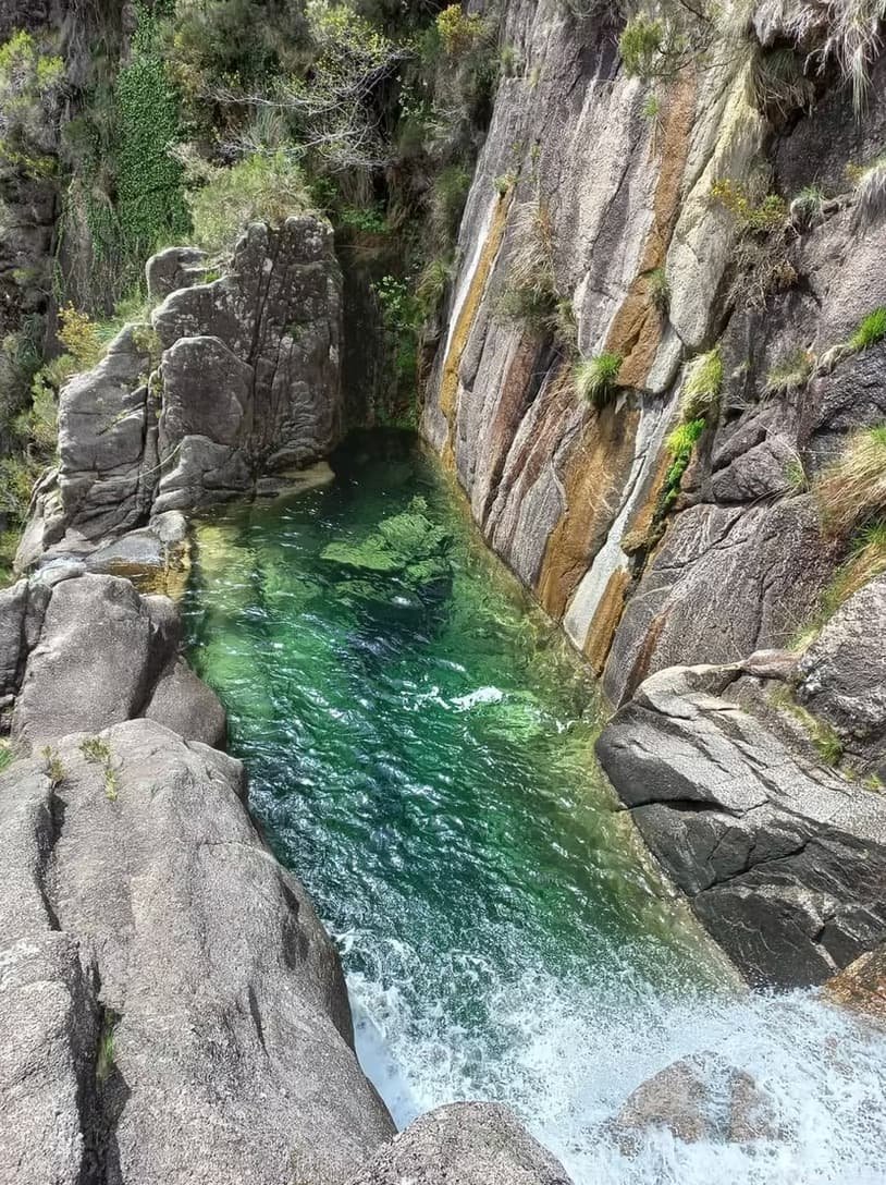 A narrow canyon with rocky walls and a small waterfall flowing into a clear green pool surrounded by large rocks and sparse vegetation.