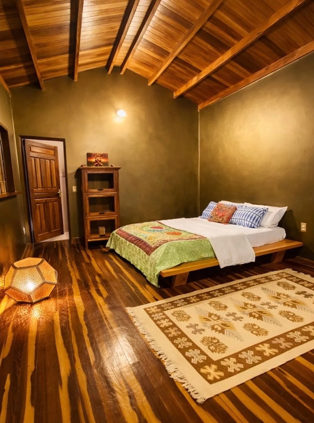 A bedroom with wooden floors and a sloped wooden ceiling. It has a bed with a green quilt, striped pillows, and a decorative pillow. There is a small wooden ladder shelf near the corner, a patterned rug on the floor, and a paper lantern-style lamp on the floor.