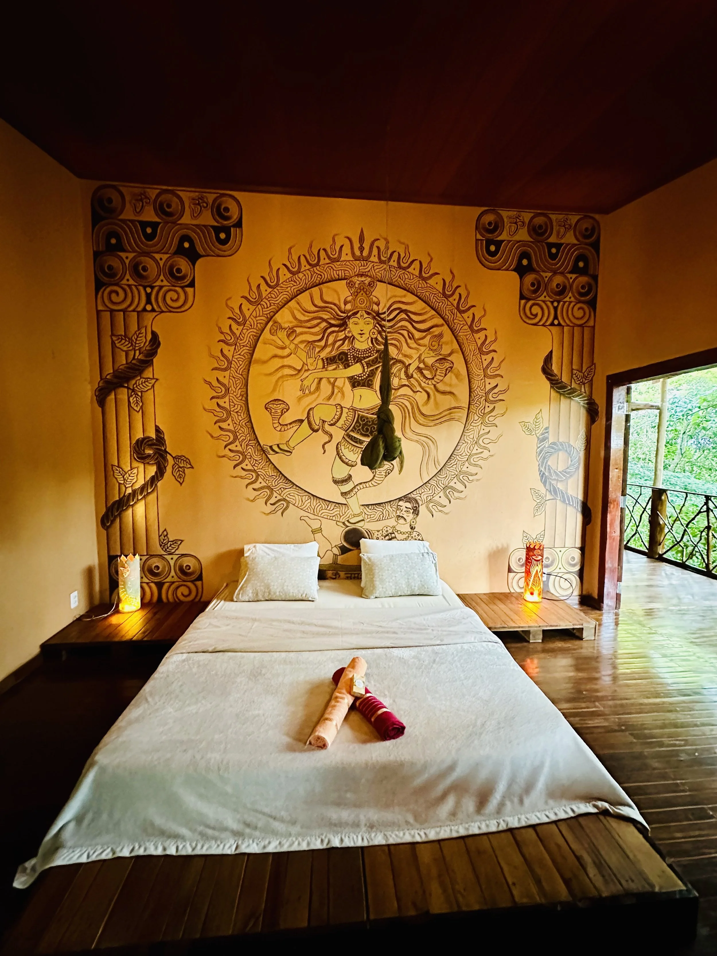 A bedroom with a decorated wall mural of a mythological figure, two pillows on the bed, and two decorated lamps on nightstands. Towels are rolled on the bed. An open balcony door shows greenery outside.