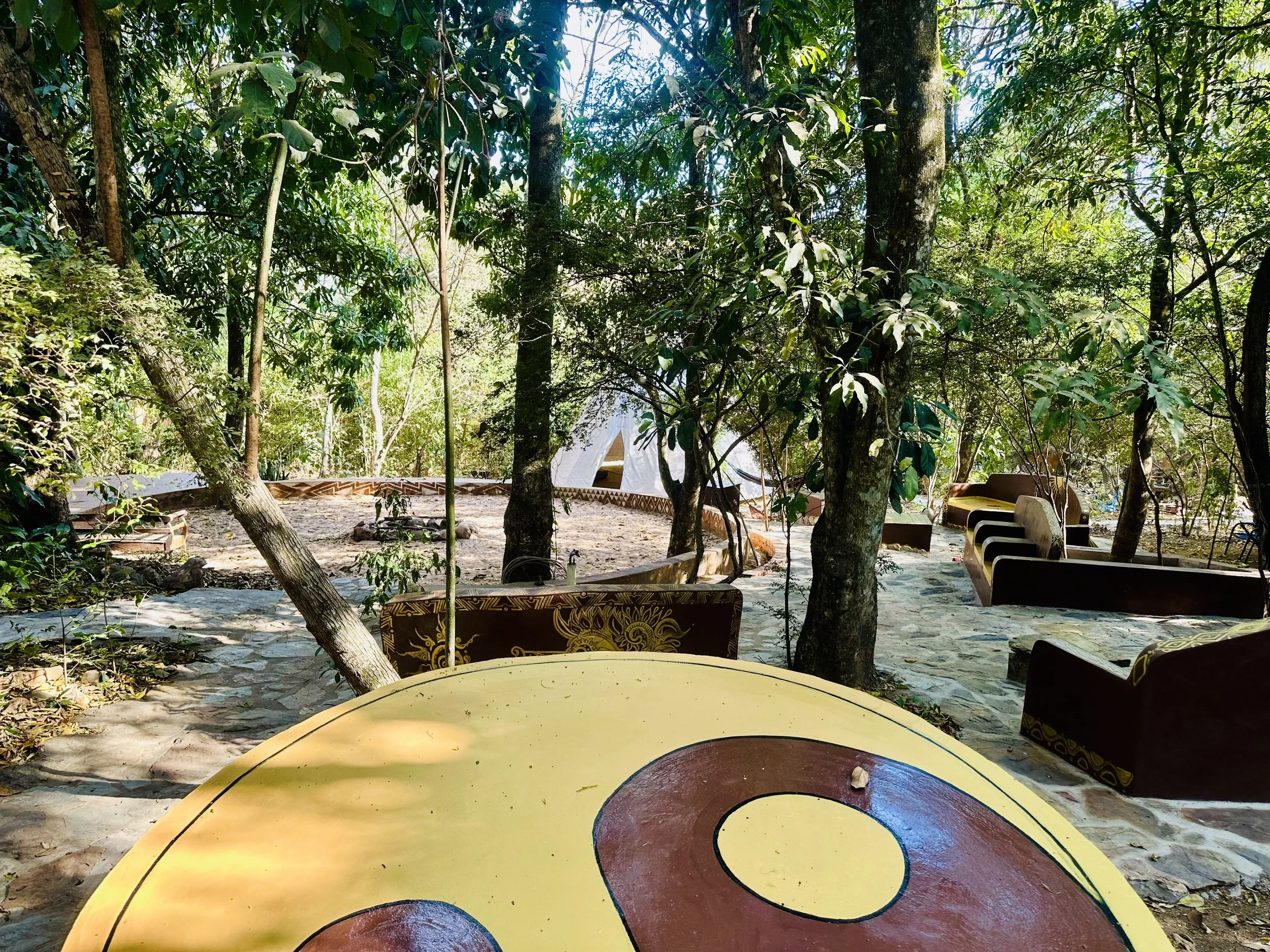 Outdoor seating area within a wooded area with trees, stone pathways, cushioned benches, and a large colorful circular table in the foreground.