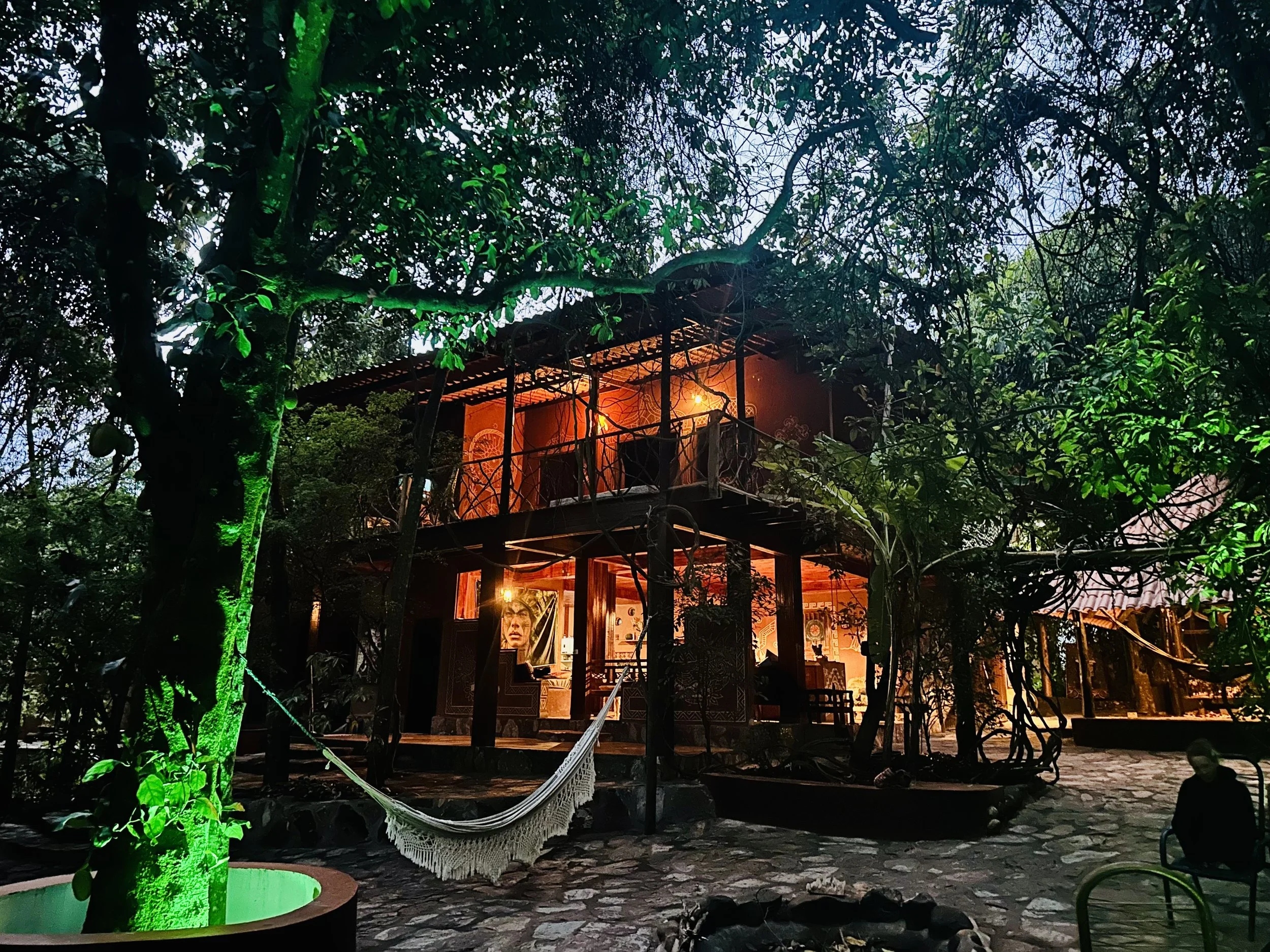 A two-story house on stilts illuminated with warm orange and yellow lighting, surrounded by trees, with a hammock hanging in the foreground and a person sitting on a chair to the right.