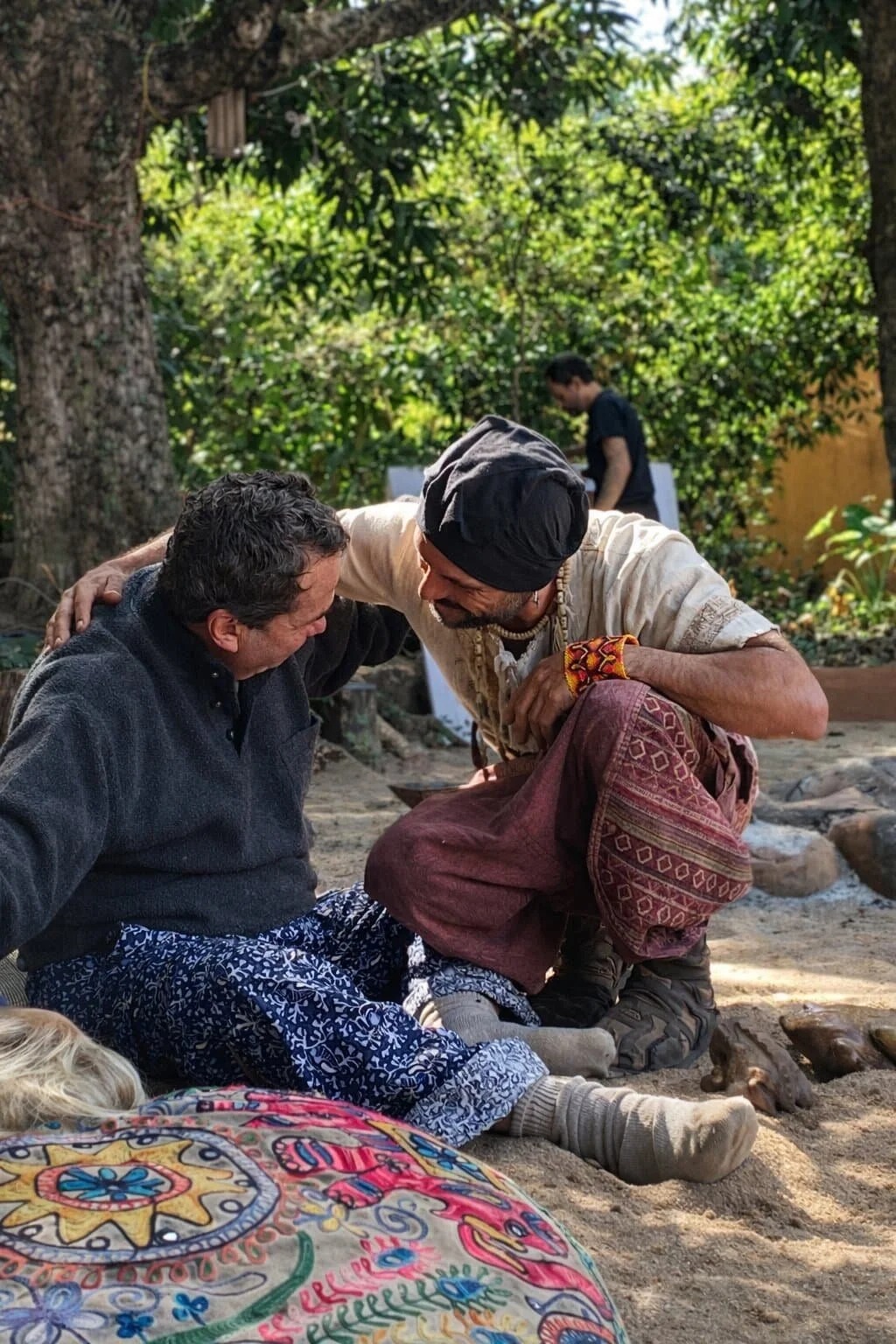 Two men are smiling and leaning towards each other in an outdoor setting, with lush greenery in the background. One is sitting on the ground wearing patterned pants and a dark jacket, and the other is kneeling, dressed in traditional attire with a black head covering and a multicolored bracelet. A third person is seen in the background, standing and looking away.