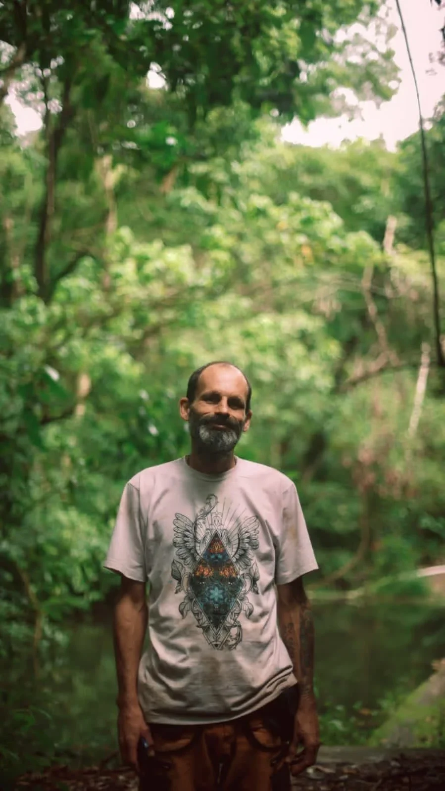 A man with a beard and tattoos, wearing a graphic t-shirt and brown pants, standing in a lush green forest near a body of water.