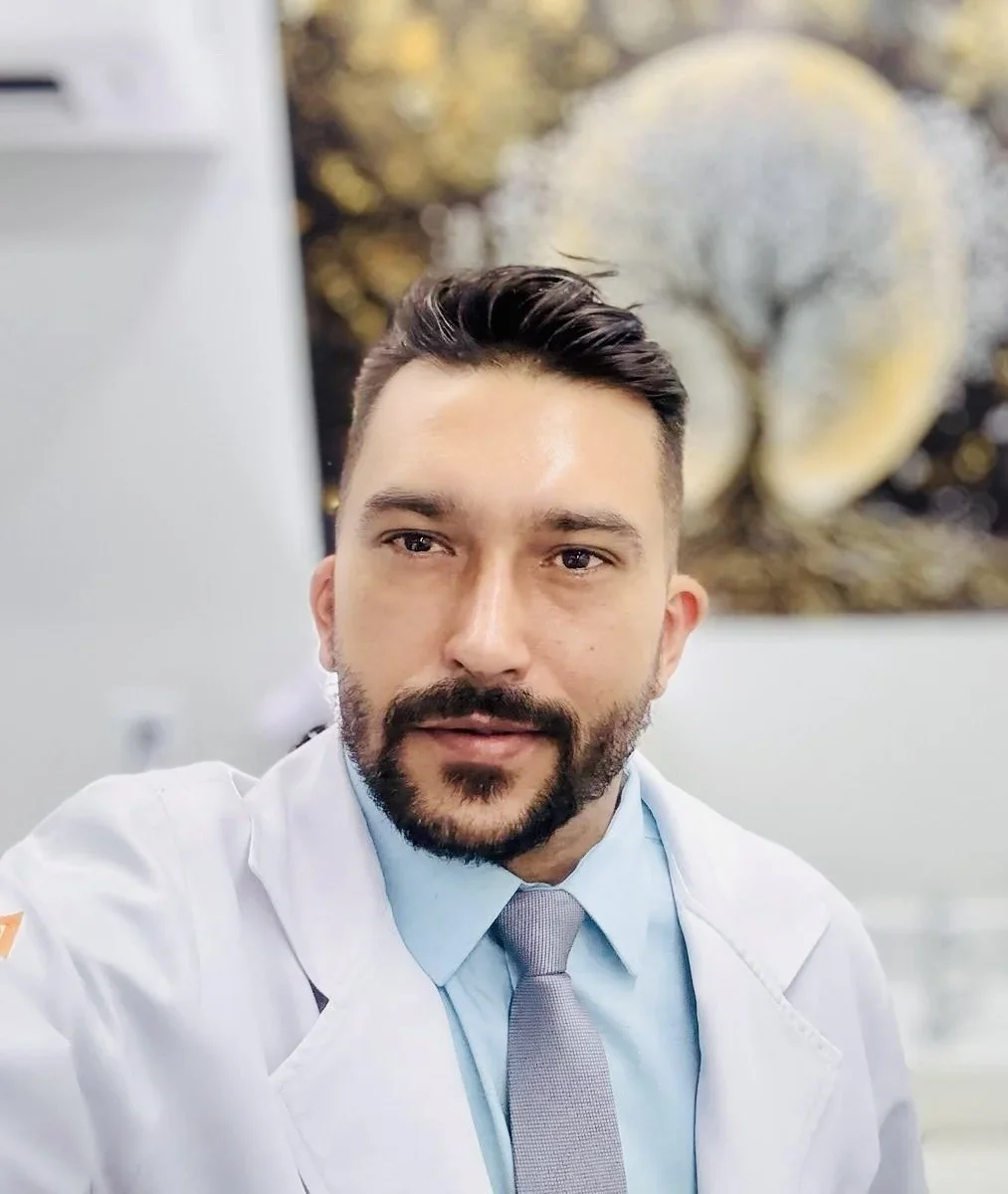 A man with dark hair and a beard wearing a white medical coat and light blue shirt with a grey tie, taking a selfie in a medical or professional setting with abstract art and a tree painting in the background.