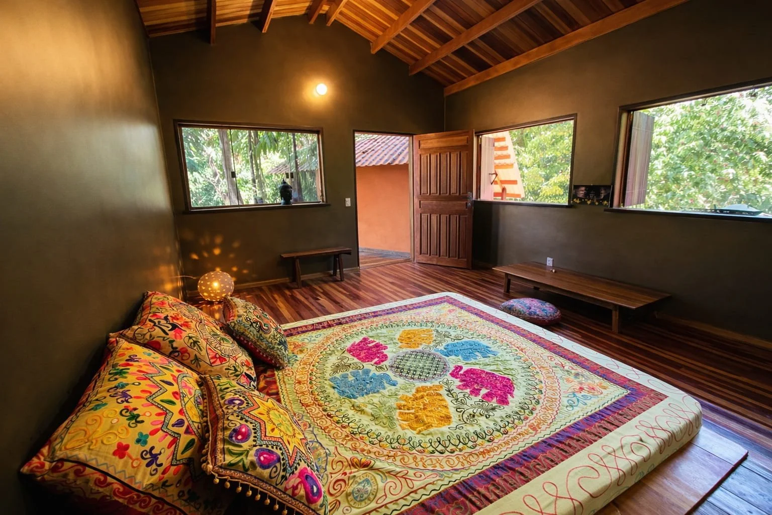 A bedroom with wooden flooring, olive green walls, large windows showing greenery outside, a wooden door, a low wooden bench, colorful embroidered pillows and a quilt on the floor, a small spherical lamp on the bed with patterned shadows, and a small cushion on the floor.