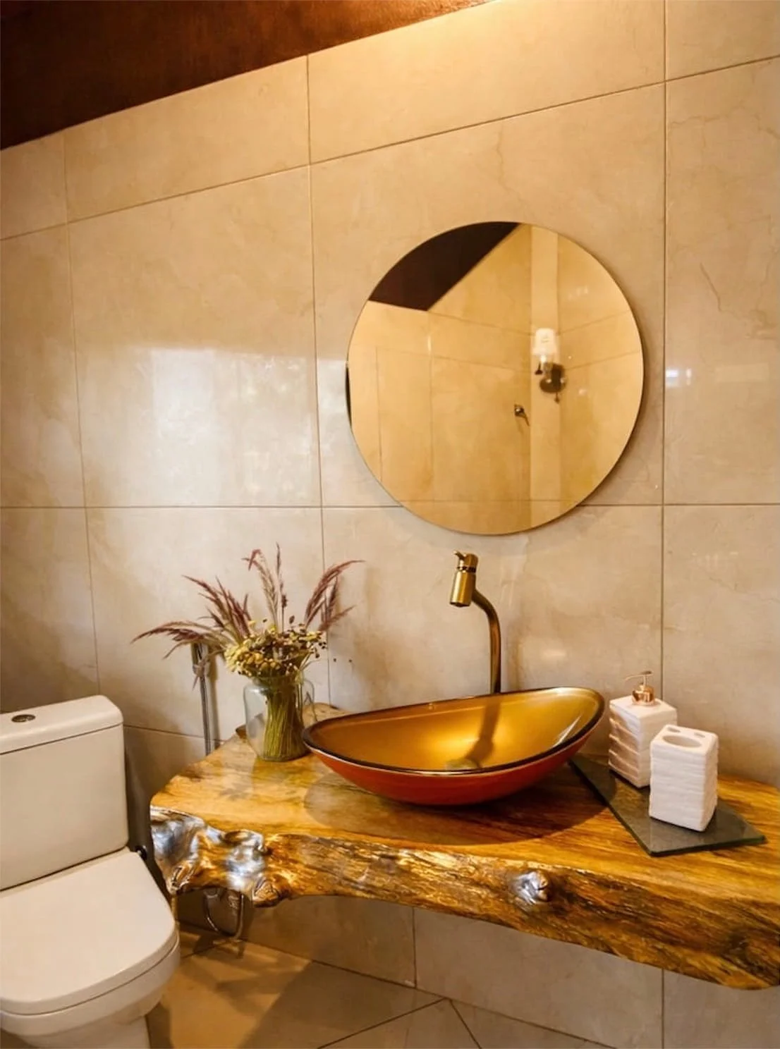 A bathroom with beige tiled walls, a round mirror, a gold and black vessel sink on a rustic wooden countertop, and decorative items including a vase with dried flowers and a tissue box.