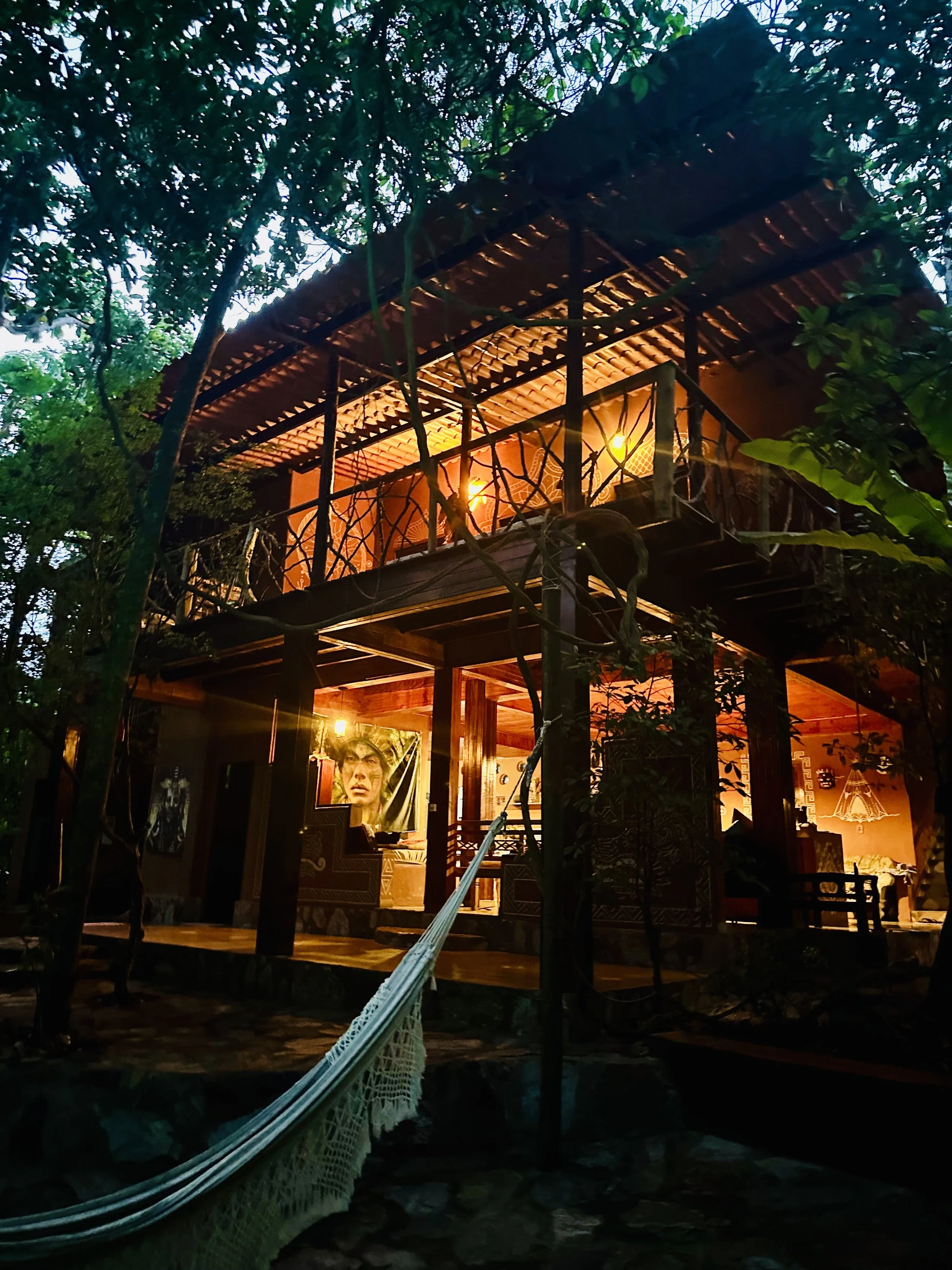 A two-story treehouse with warm lighting, surrounded by trees, with a hammock hanging nearby.
