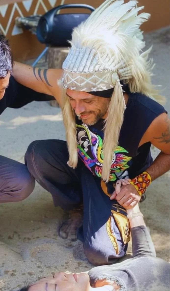 Two men appear to be helping a woman who is laying on the sand, possibly unconscious. One man is dressed in a black hoodie and khaki pants, while the other is wearing a colorful feathered headdress, black outfit with a colorful design, and a beaded bracelet. They are supporting her head and shoulder as she lies on the ground.