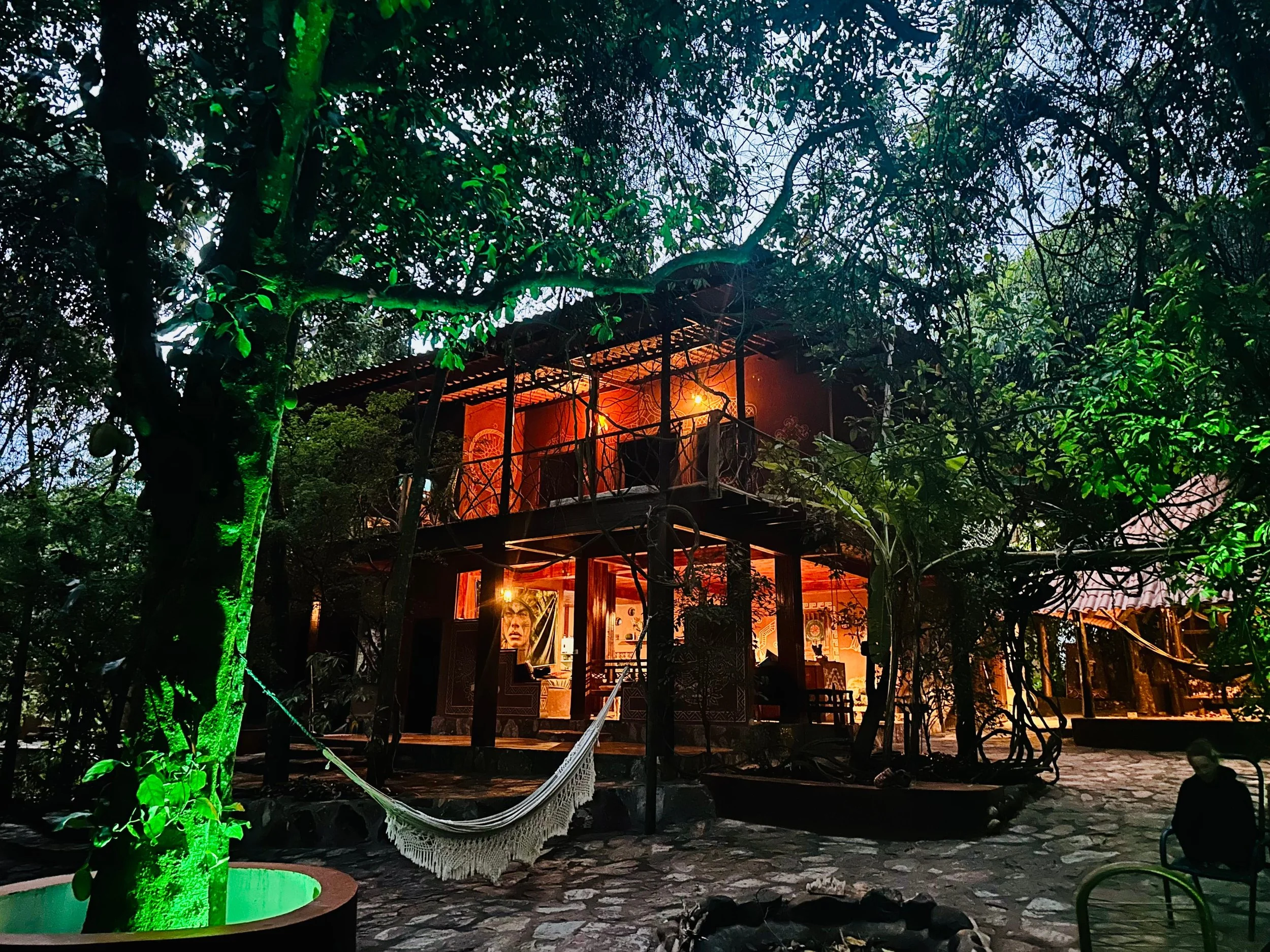 A two-story wooden house with warm interior lighting, surrounded by trees, with a hammock hanging in the front yard and a person sitting on a chair outside.