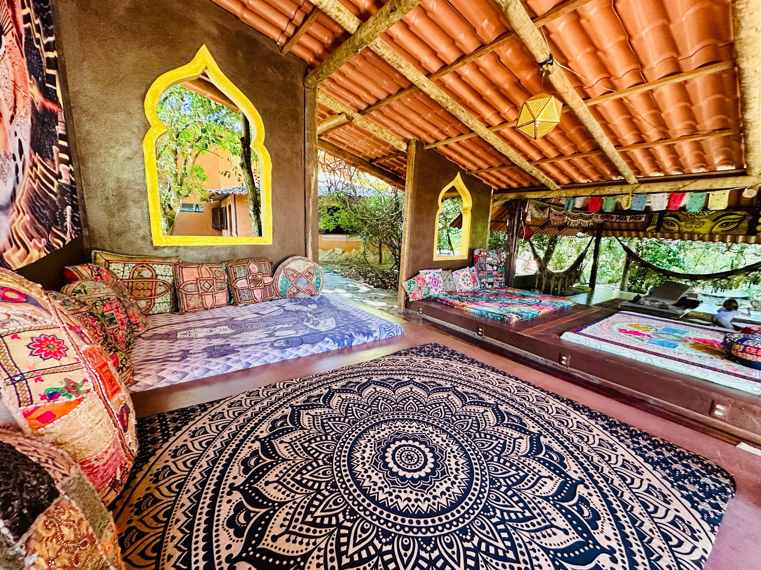 Open-air lounge with colorful patterned cushions on built-in beds, traditional Indian-style arched windows with yellow trim, decorative textiles, a large black and white mandala rug, hammock, and lush greenery outside, under a terracotta tiled roof with bamboo beams.