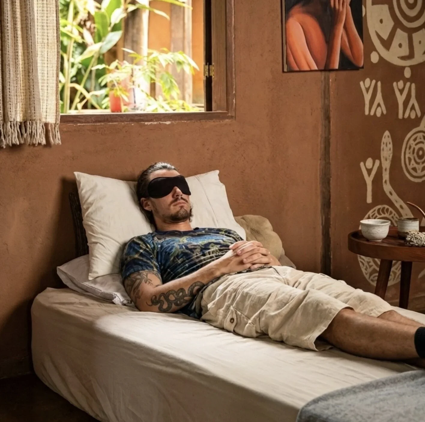 A man with tattoos on his arm, wearing a T-shirt and beige shorts, is lying on a bed with his hands clasped on his stomach. He is wearing a black sleep mask and resting with his eyes closed. The room has orange walls and a window open to green plants outside. There is a small wooden table next to the bed with white bowls and a decorative item.