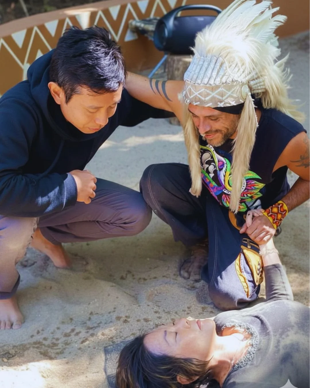 Two men support a woman lying on the ground as she appears to be in distress. One man wears a traditional indigenous headdress and colorful attire; the other man wears a black hoodie. They are on a sandy surface, possibly outdoors.