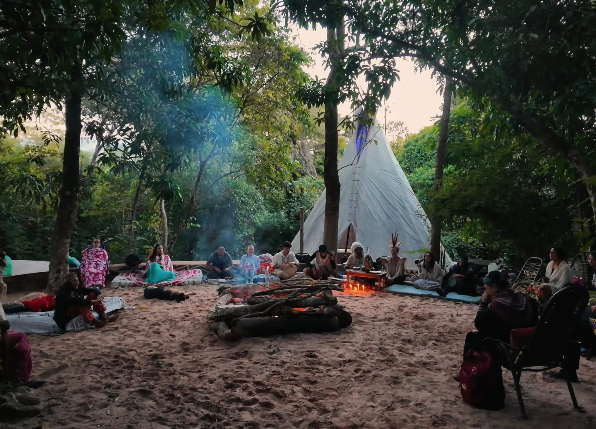 Group of people sitting around a campfire in a forest, with a tipi tent and trees in the background.